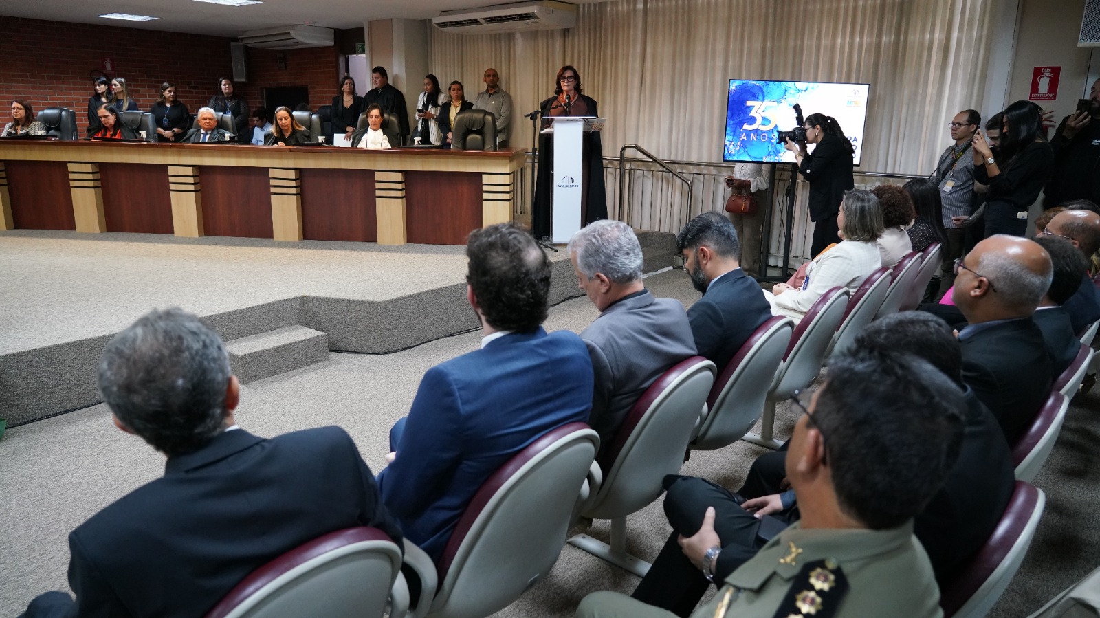 imagem aberta de parte do pleno do TJTO, ao fundo a desembargadora Etelvina em pé no pulpito, de roupa marrom e toga escura, fatando ao microfone