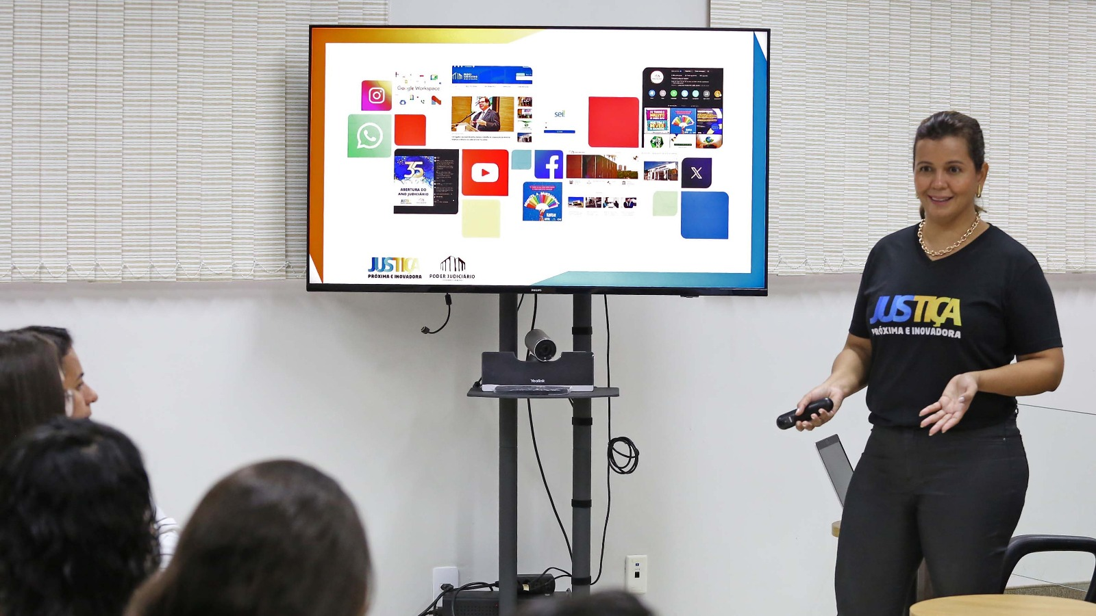 Diretora Kezia Reis diante de uma tela de TV com ícones de redes sociais, sites e ferramentas de comunicação do TJTO