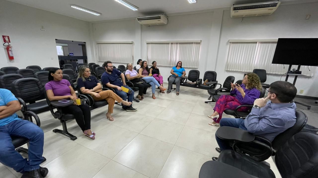 Equipe do Napsi e servidores reunidos em uma roda de conversa; eles estão em uma sala, sentados em cadeiras pretas; a sala possui paresdes e cortinas brancas, dois aparelhos de ar condicionado e um monitor