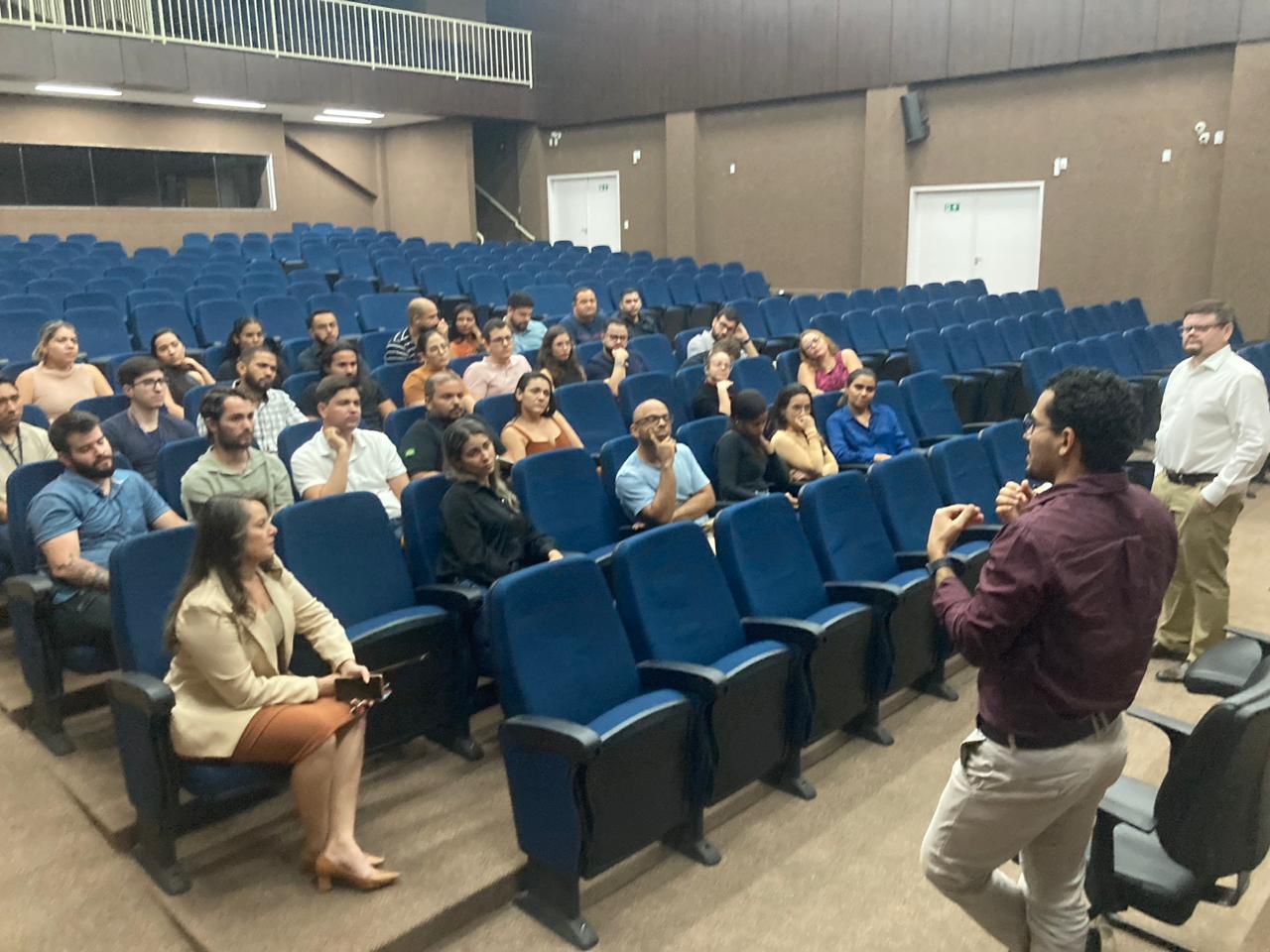 Servidores sentados em cadeiras azuis, no auditório; em primeiro plano os dois integrantes da equipe do Napsi conversam com os servidores