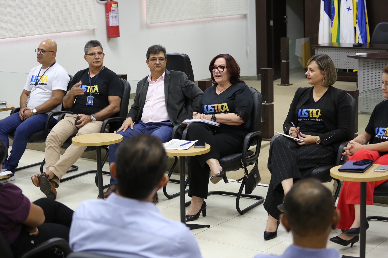 Desembargadora Etelvina, de roupa preta com a logo da Justiça Mais Próxima, está sentada, ao lado do juiz Jorge Amâncio e sua equipe, conversando com os servidores 