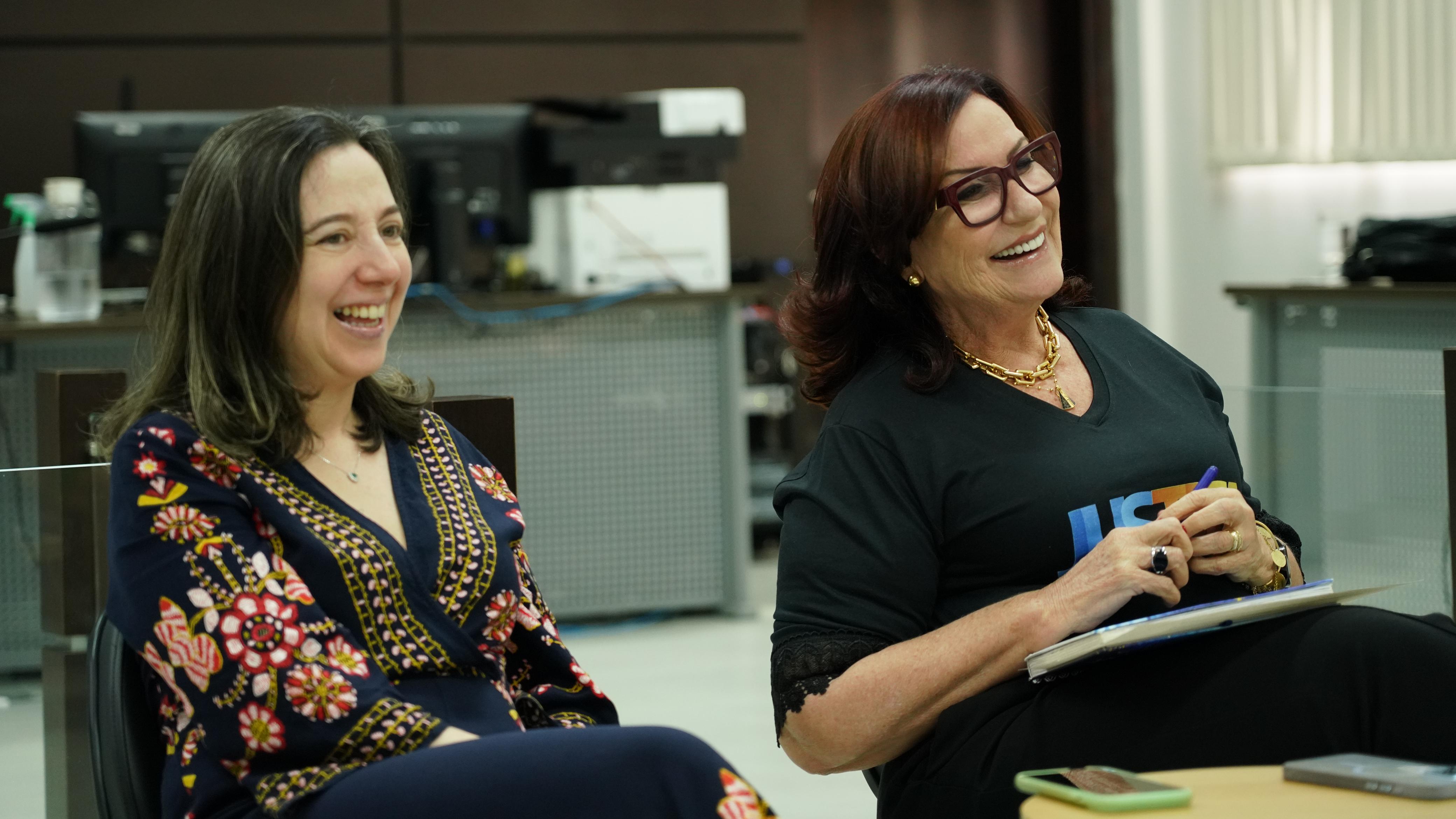 Desembargadora Etelvina (à direita), de camiseta preta com a Logo do Justiça Mais Próxima, em momento de descontração ao lado da juíza Emanuela da Cunha Gomes, uma mulher branca de cabelos castalhos que está sorrindo e usa ropa azul-marinho com estampas florais em tons de vermelho, amarelo e branco 