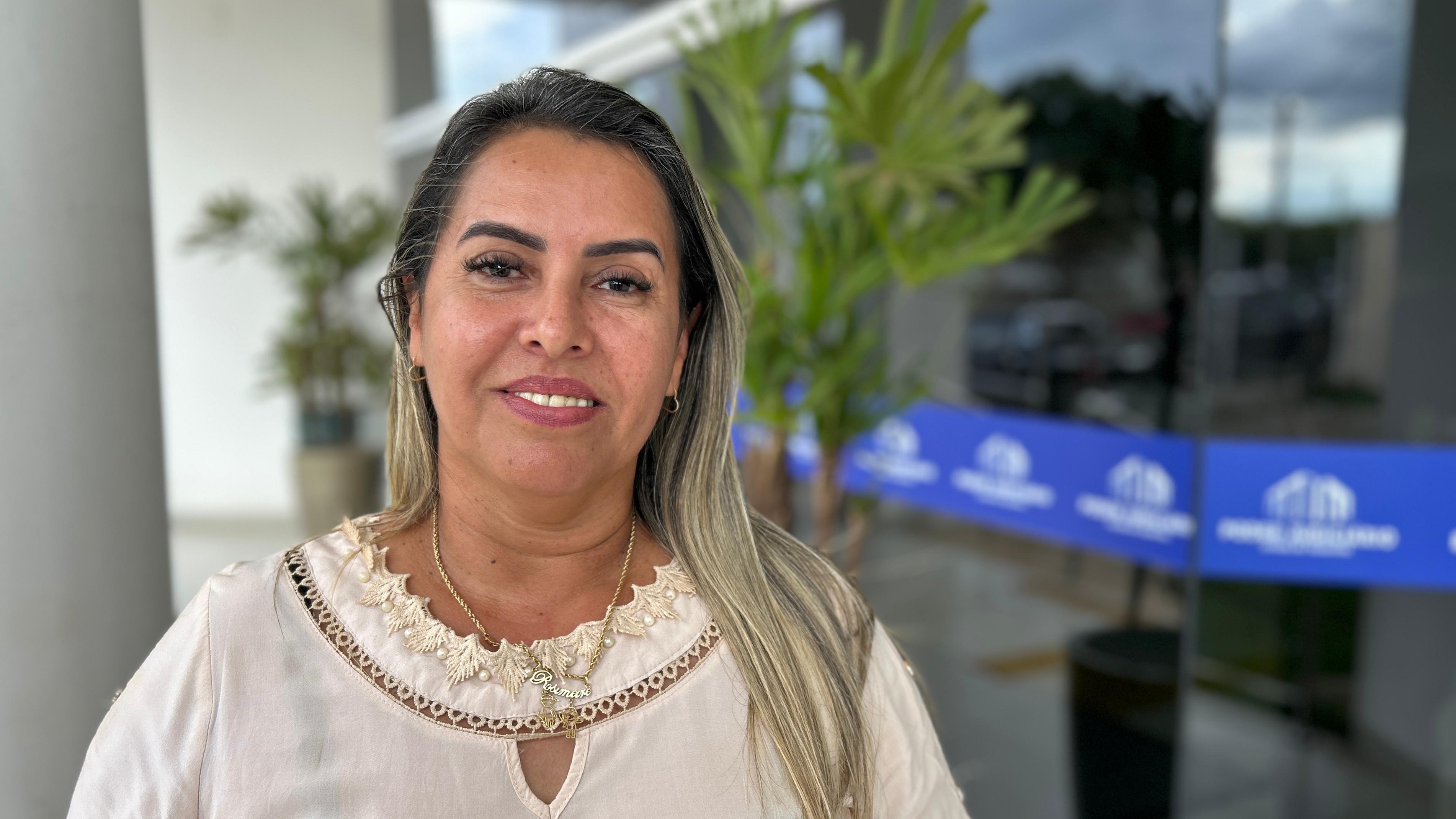 Rosimeire é uma mulher branca, de cabelos louros. Ela usa blusa branca e está sorrindo para a foto