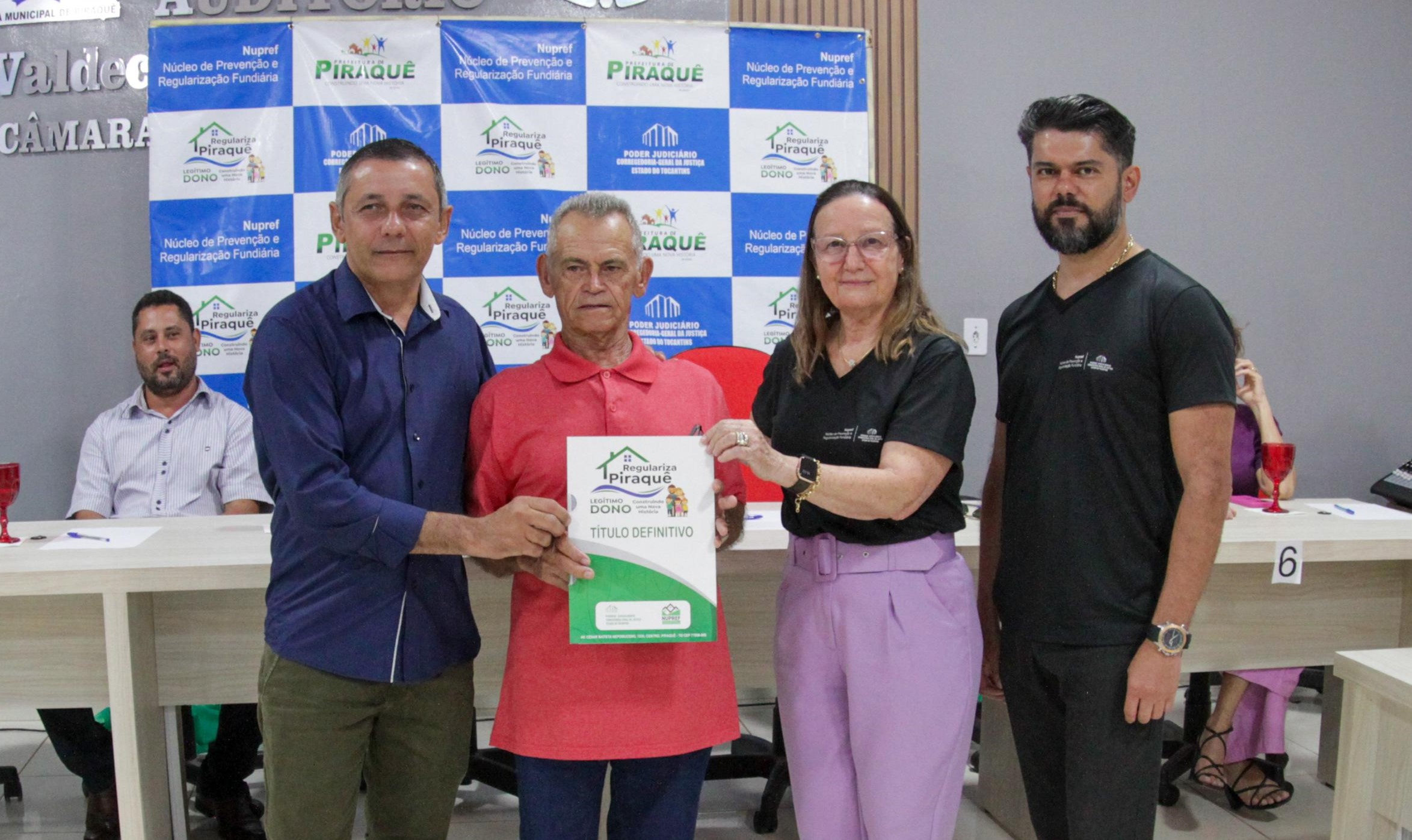 Quatro pessoas pousando para a foto e três deles estão segurando o documento de regularização fundiária na cor branca e verde, o homem do lado esquerdo veste uma camiseta social na cor azul escura e calça verde musgo, o homem do lado dele veste uma camiseta polo de cor vermelha e uma calça jeans azul escura, a mulher ao lado veste uma blusa de cor preta e uma calça lilás, já o homem que não segura o papel veste uma camiseta preta e uma calça jeans preta, ao fundo tem um homem sentada de camiseta social azul e uma mesa na cor branca