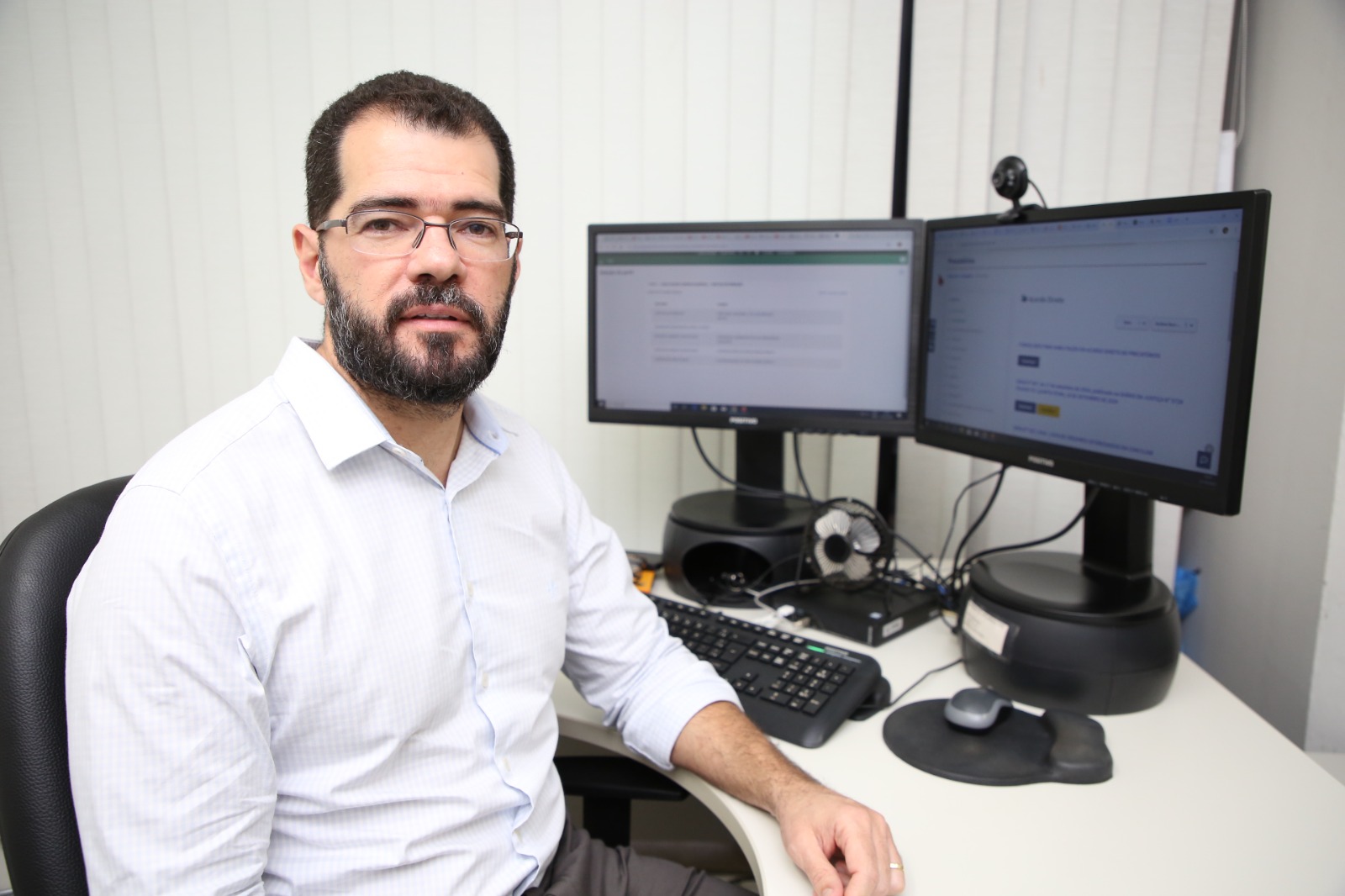 Juiz Manuel de Farias, um homem de pele clara, cabelos castanhp escuro e barba grisalha; ele usa óculos e está de camisa branca, sentado em uma cadeira, ao lado de uma mesa com dois monitores, teclado e mouse.