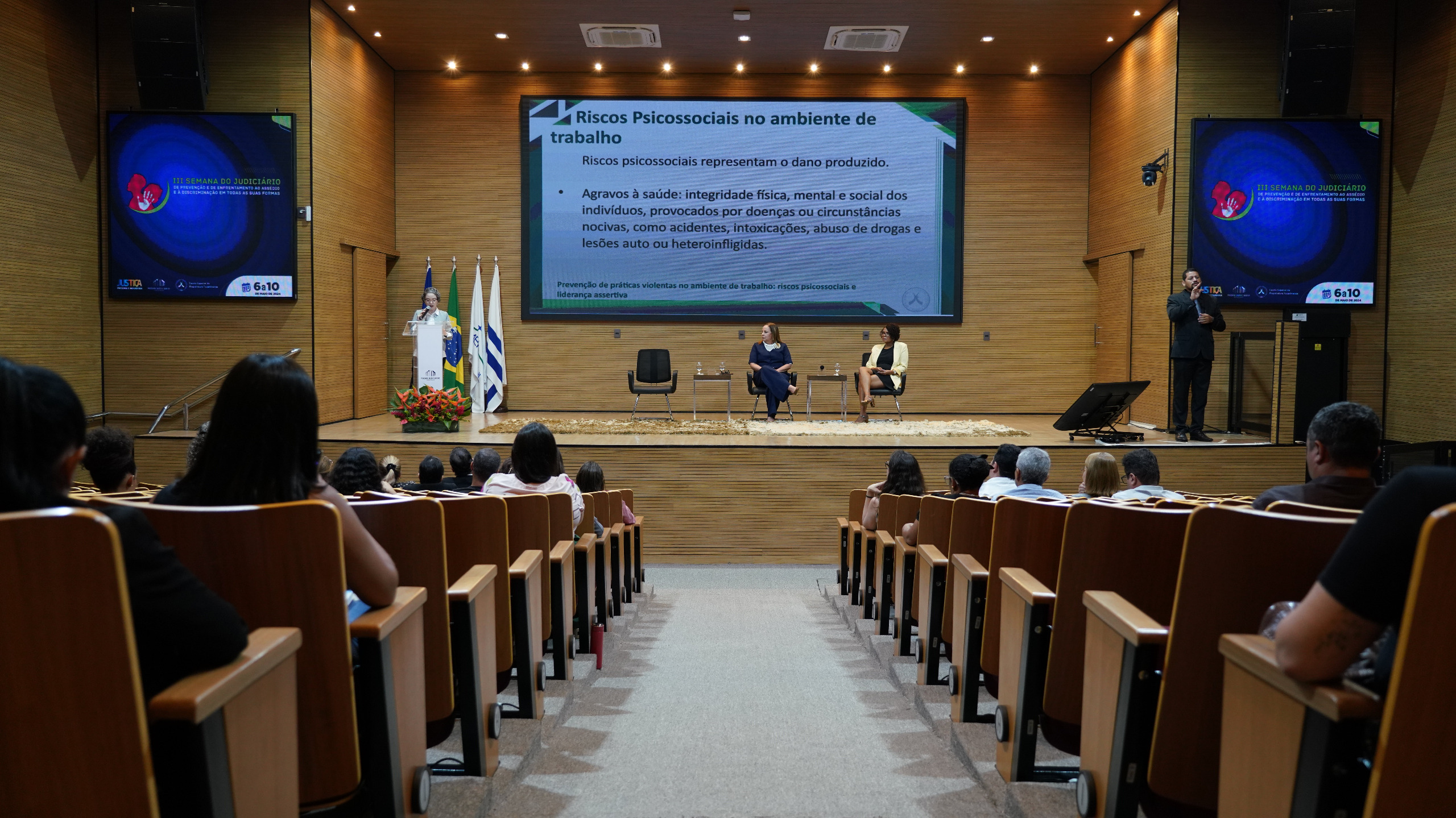 Auditório do Tribunal de Justiça do Tocantins, com várias cadeiras de madeira e algumas pessoas assistindo a palestra de costas para a foto, no final da foto tem um telão com o slide da apresentação da palestra, tem três cadeiras pretas, mas apenas duas mulheres estão sentadas, uma estar com vestido preto e blazer amarelo claro e a outra estar sentada ao lado com macacão azul escuro, e em pé falando no púlpito estar uma outra mulher  com blazer cinza.