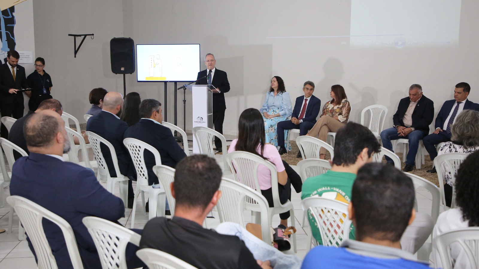 Homem branco e cabelos grisalhos fala ao microfone em púlpito, posicionado em frente a tela em auditório composto por homens e mulheres sentados em cadeiras plásticas brancas. As paredes são brancas. 