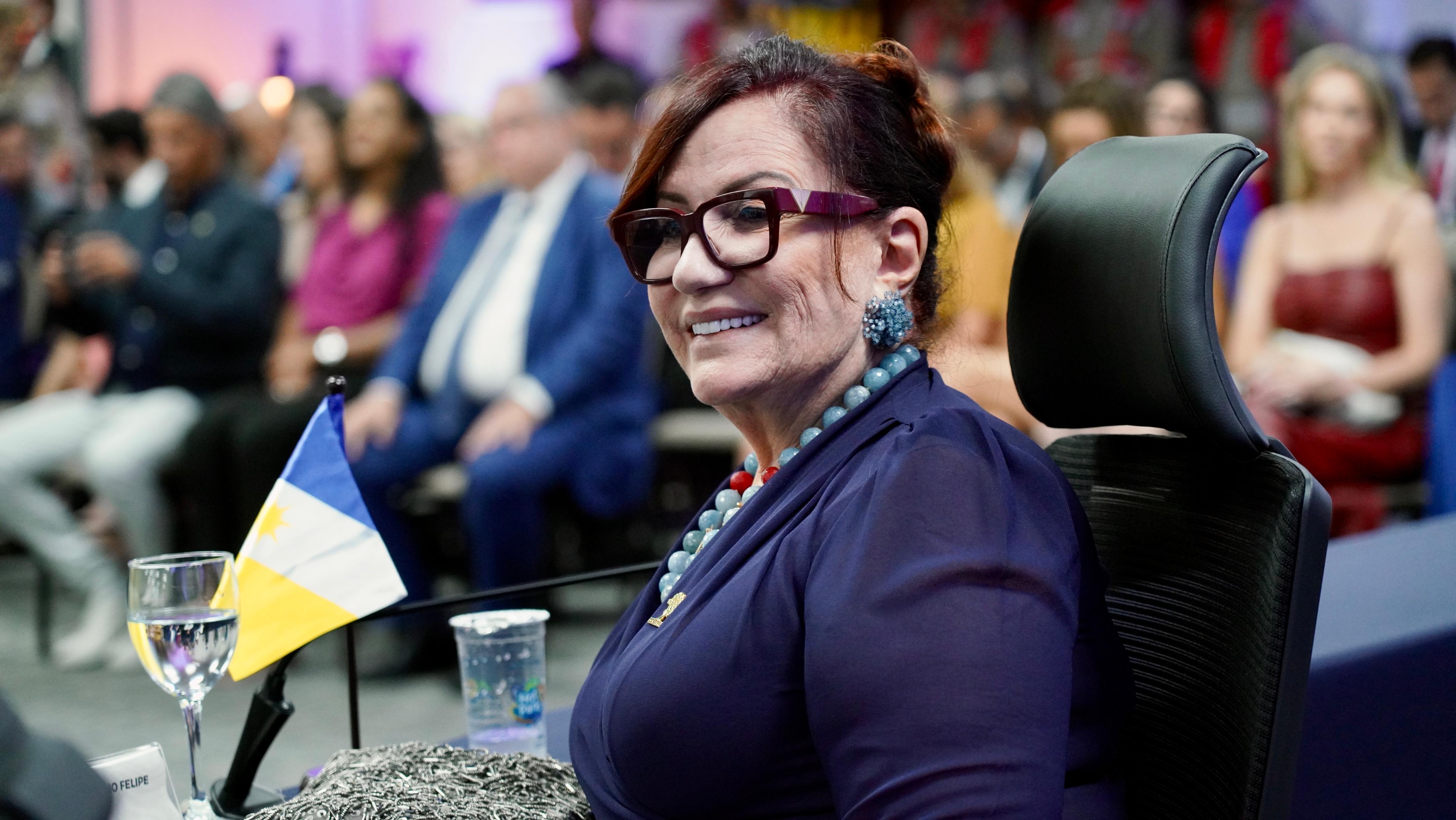 Imagem mostra uma mulher branca com terno azul escuro, óculos de grau e cabelo preso sentada; à frente uma mesa com a bandeira do Tocantins