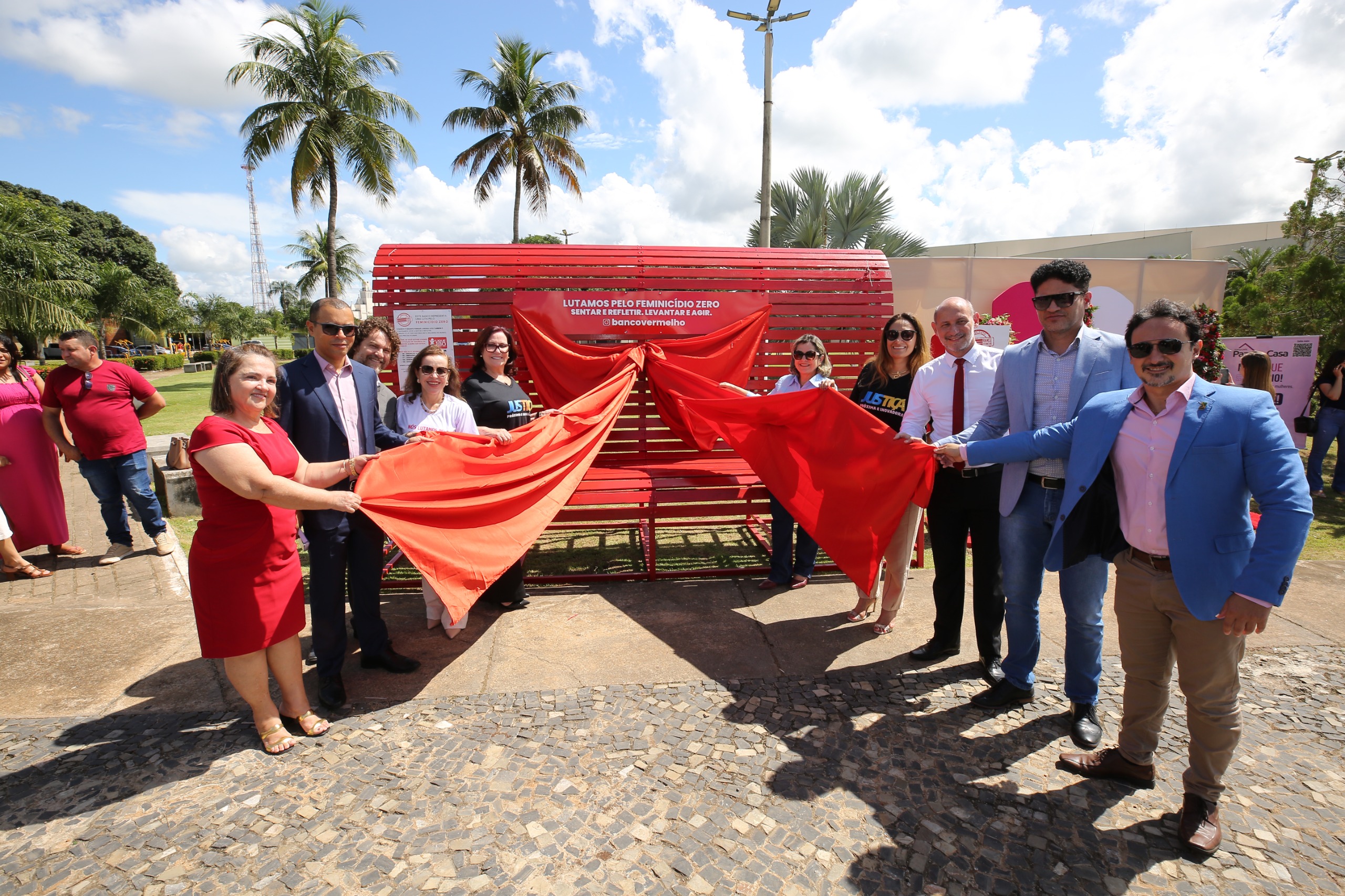 Fotografia colorida onde várias pessoas estão de frente umas para as outras e ao lado de um grande banco vermelho. Elas estão segurando um laço que cobre todo o banco e se preparam para desfaze-lo. Eles estão em um espaço aberto com palmeiras altas no fundo. 