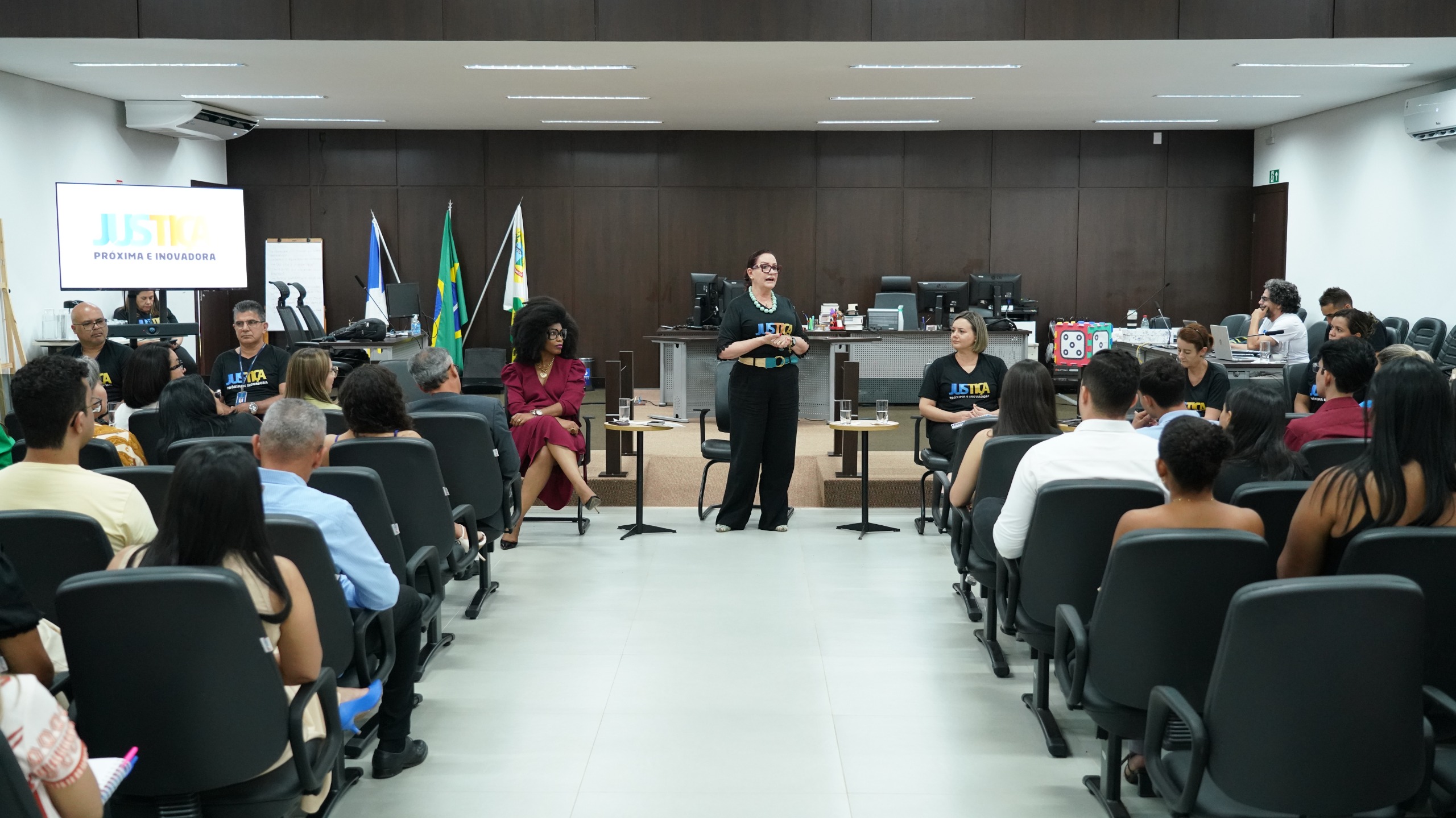 Imagem mostra um auditório com várias pessoas sentadas em cadeiras pretas; à frente uma mulher de preto fala ao microfone e ao lado estão outras pessoas sentadas