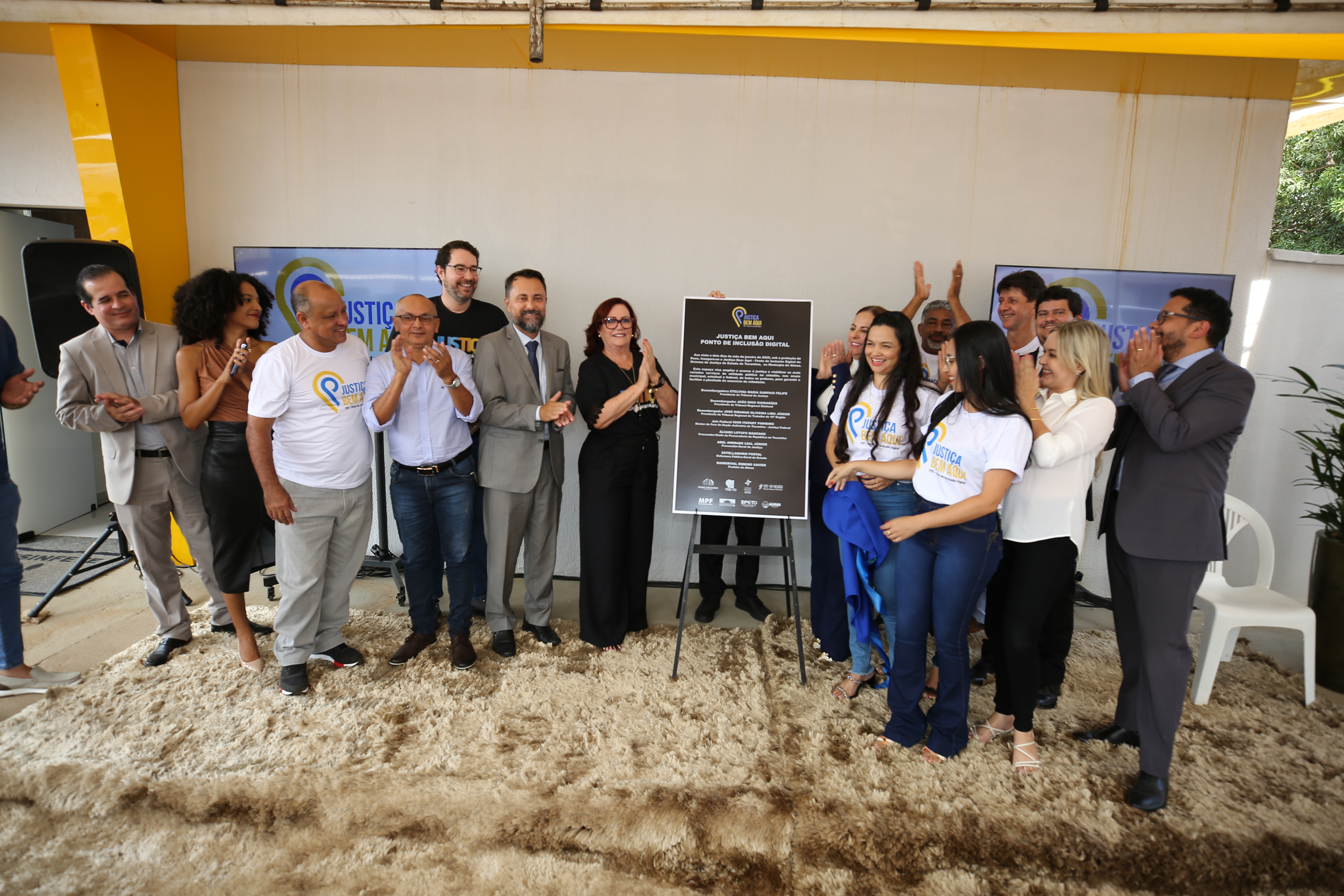 Fotografia coloprida onde várias pessoas estão batendo palmas ao lado de uma placa preta com os dizeres "Ponto de Inclusão Digital". Ao fundo, tem uma parede branca com uma borda amarela e abaixo um tapete felpudo amarelo queimado 