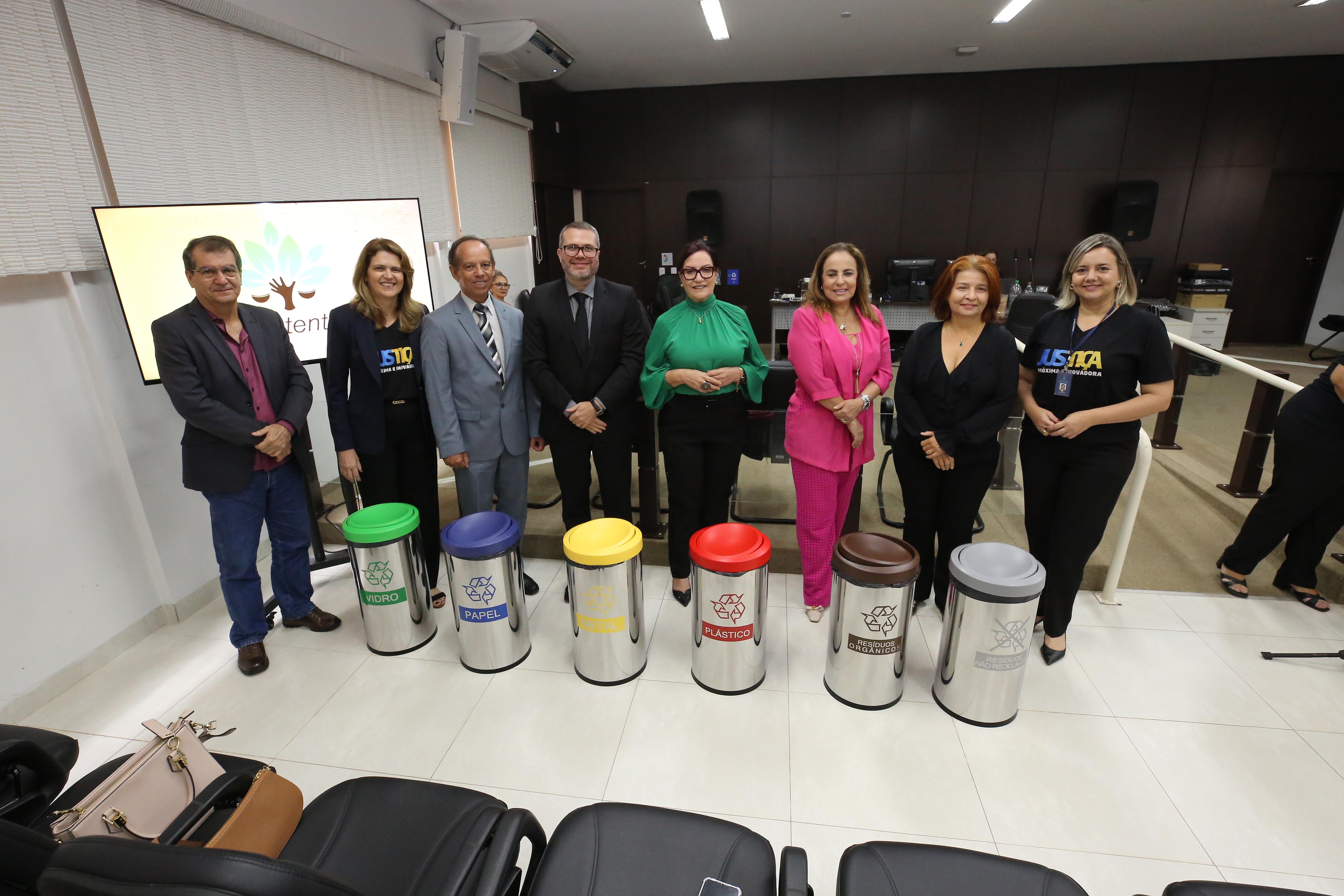Fotografia colorida onde cinco mulheres e três homens estão em pé posando para foto na frente de lixeiras de reciclagem. Eles estão em uma sala branca com uma parede preta ao fundo, e atrás deles uma televisão está ligada