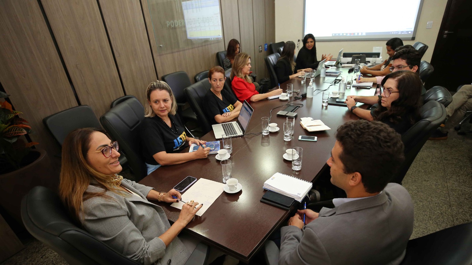 Fotogrfia colorida onde várias pessoas estão sentadas em volta de uma grande mesa retangular de madeira escura, em cadeiras almofadadas pretas em uma sala com predes de madeira escura. Ao fundo tem uma tela fixa na parede ligada 