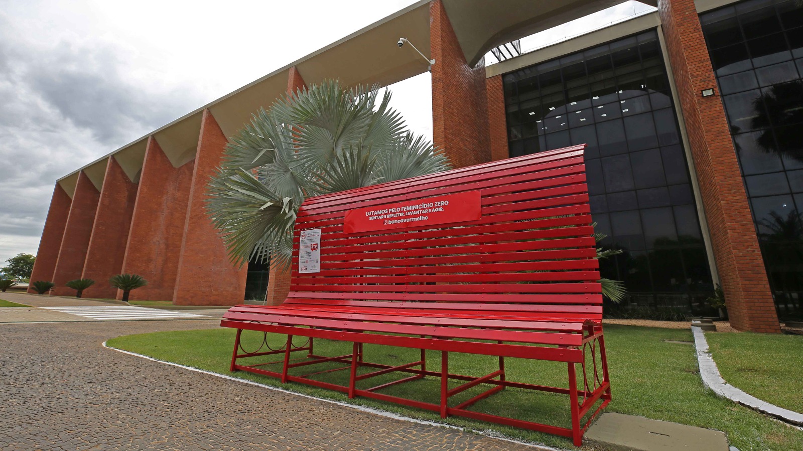 Banco Vermelho gigante instalado em praça pública com gramado e palmeiras ao redor
