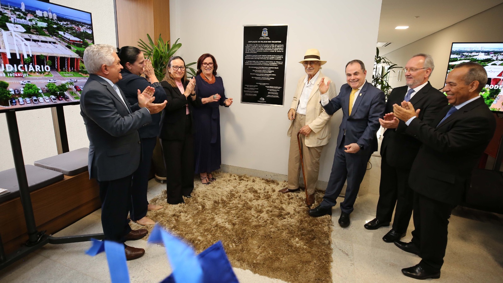 Homens e mulheres em pé aplaudindo durante descerramento de placa de inauguração de obra em parede branca. Duas telas de tv, uma de cada mostra a obra de ampliação do TJTO, no chão, um tapete marrom.