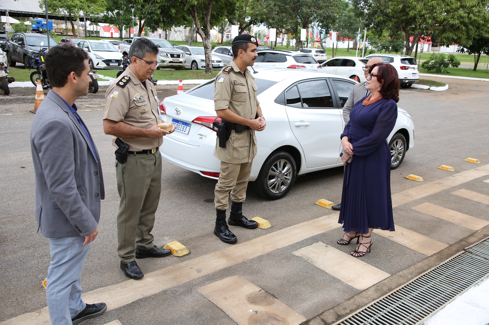 Fotografia colorida onde a presidente do tribunal de Justiça está de pé ao lado de um carro branco junto de dois policiais e um homem com terno cinza. Eles estão em uma área externa, de pé sobre uma rua com árvores ao fundo  