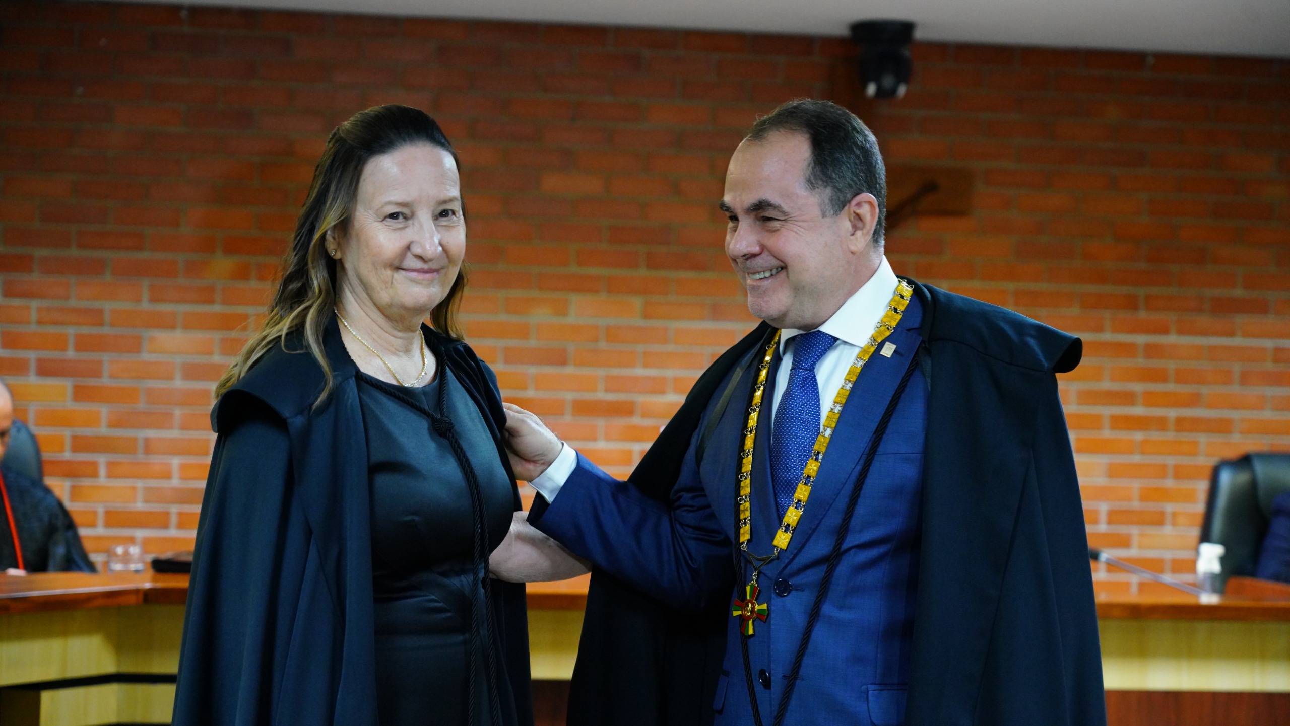 Mulher branca de cabelos lisos soltos usando toga é cumprimentada por homem branco de terno azul e toga