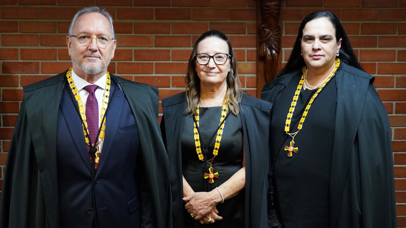 Homem branco de cabelos grisalhos e óculos de grau, trajando terno azul ao lado de duas mulheres brancas de roupas pretas. Todos usam toga e medalhas condecorativas
