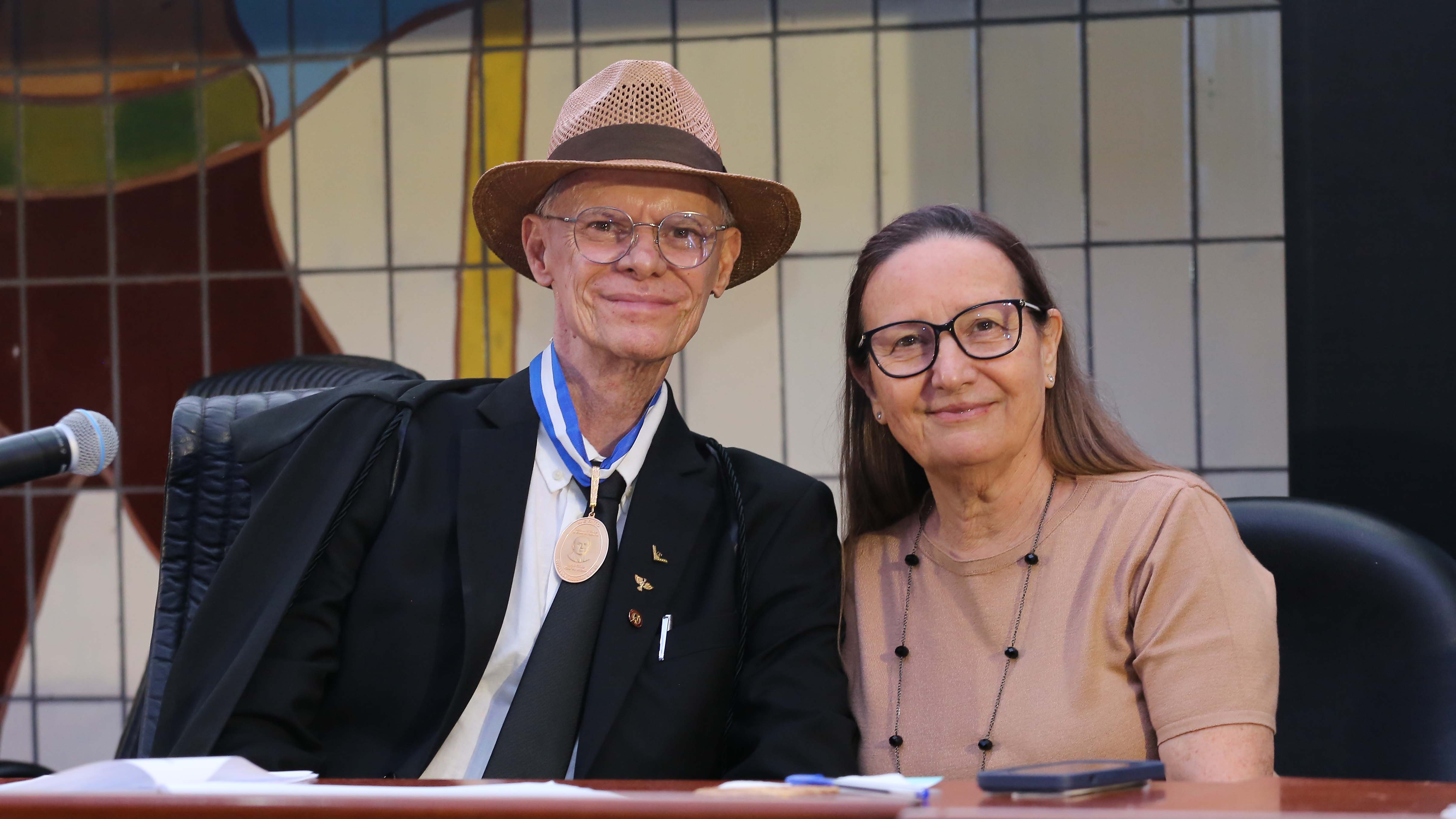 Desembargadora Maysa (à direita), de blusa bege, colar de corrente e bolas pretas, e óculos,  posa ao lado do novo presidente do TCETO, de camisa branca, blazer e gravata preta, beca escura; ele usa medalha no pescoço, óculos e chapel