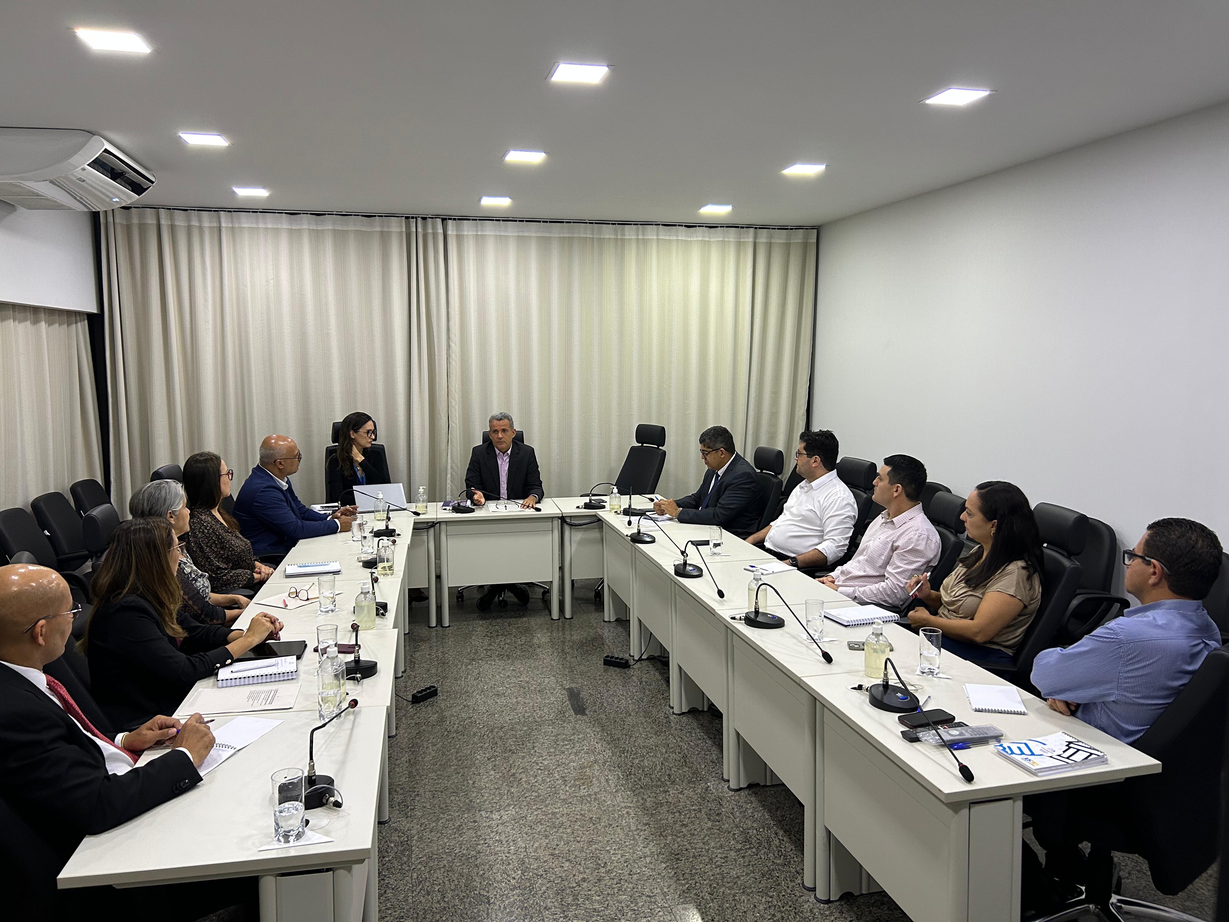 Homens e mulheres sentados em mesa branca em formato U, em sala de paredes brancas e persiana bege