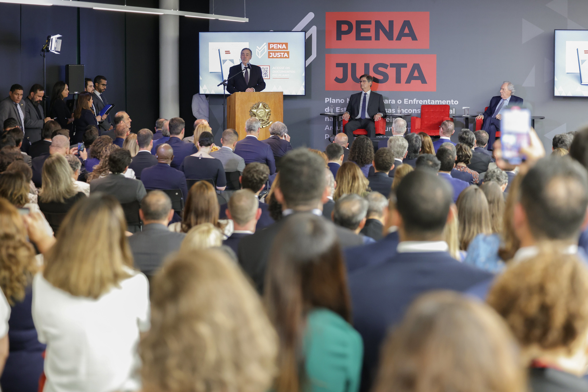 Homem de terno preto fala ao púlpito para plateia de auditório. Ao fundo, parede preta, com dizeres em vermelho sobre lançamento de programa sobre cumprimento de pena