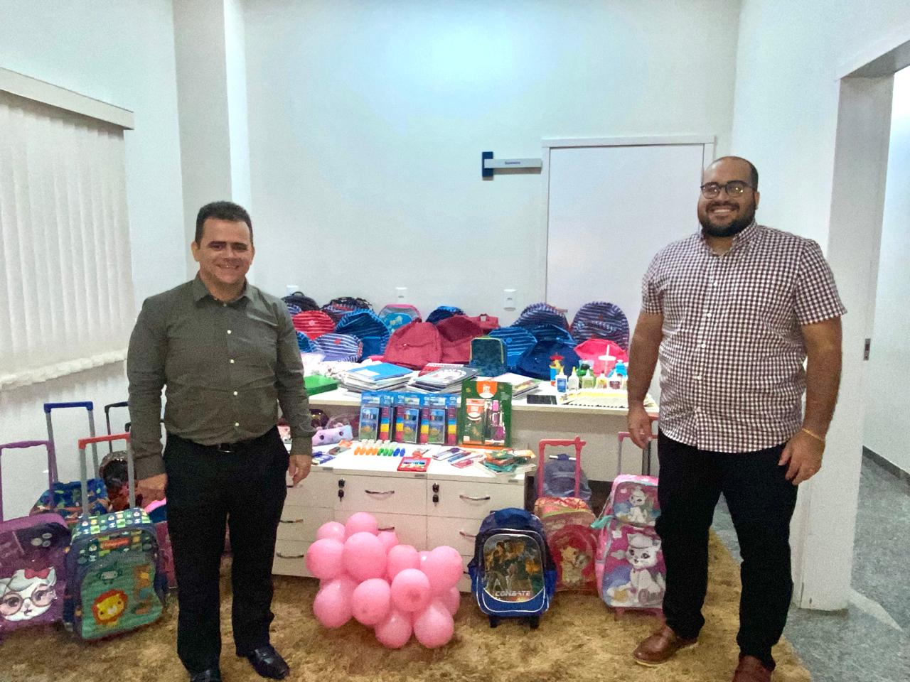 Homem de calça preta e camisa verde oliva no canto esquerdo da sala, no canto direito, um homem alto, com calça preta e camisa de manga curta xadrez clara. A sala de paredes brancas com um tapete bege tem uma mesa e abriga diversos materiais escolares arrecadados para doação