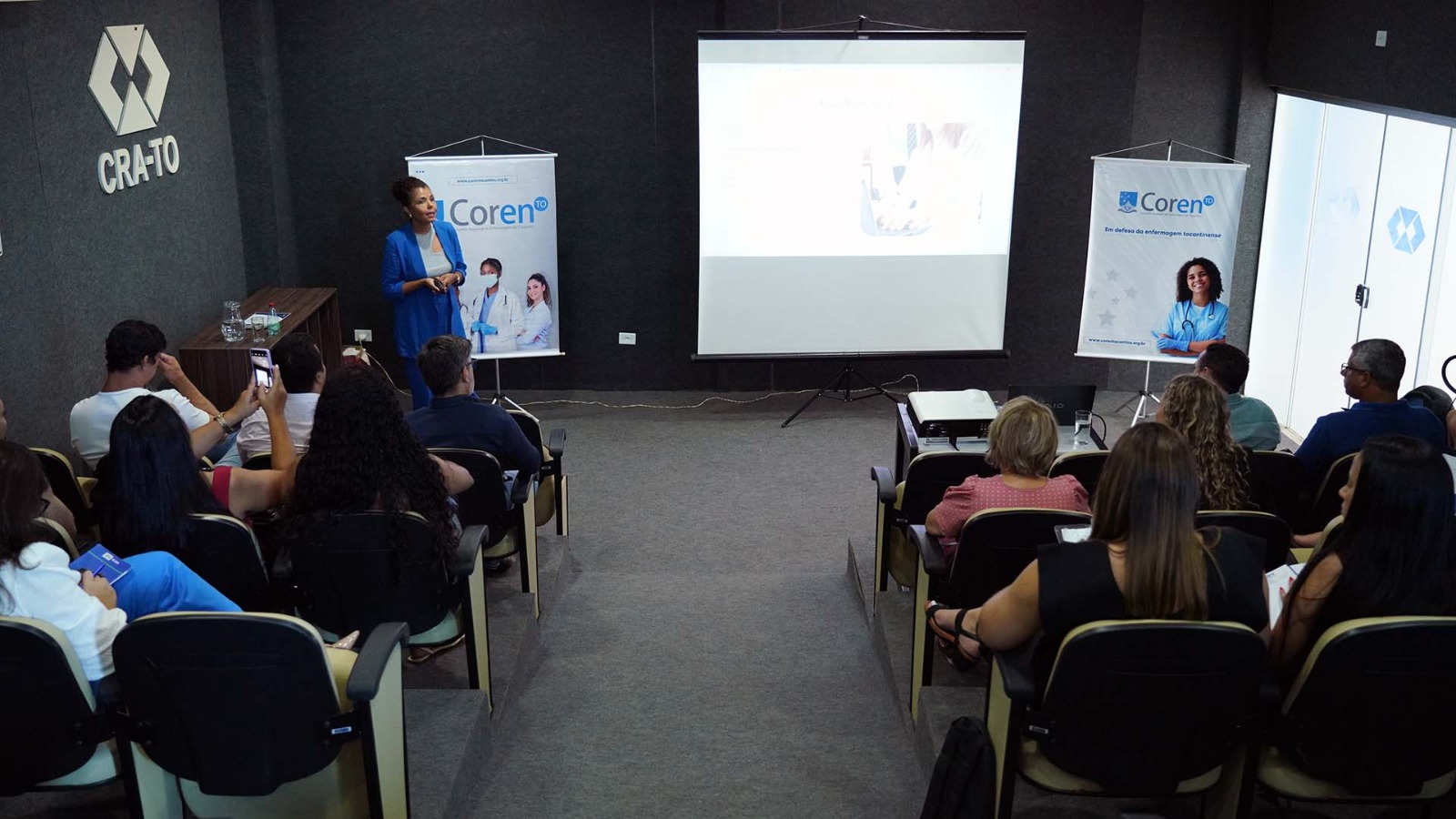 Mulher negra de camisa cinza e terno azul fala para público em auditório de paredes e piso em tons cinza escuro. Ao fundo, telão e banners em tons brancos e azuis claros sobre tema assédio