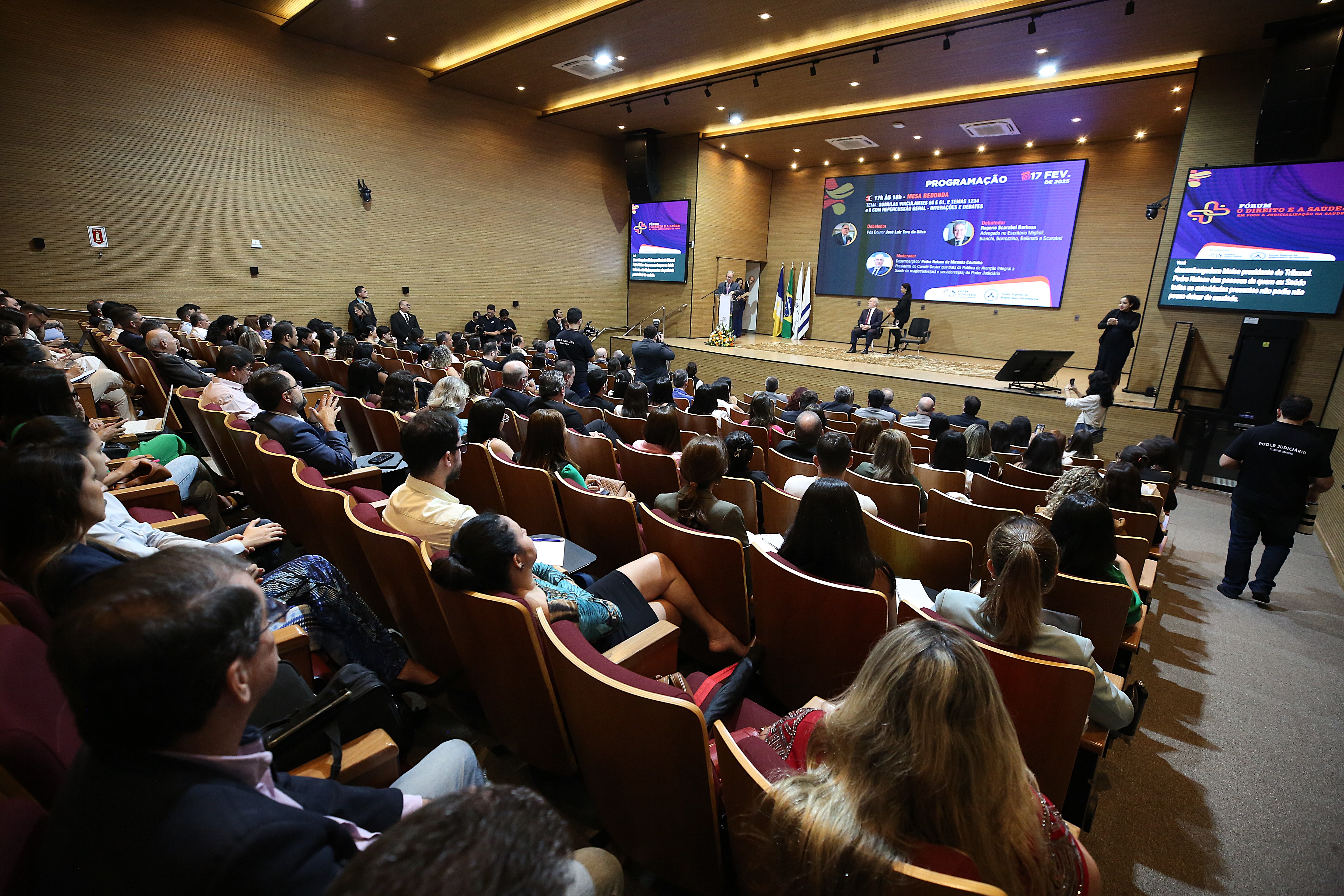 Homens e mulheres sentados em auditório lotado assistem palestra em telão ao fundo de palco. O espaço tem paredes e teto em acabamento em madeira