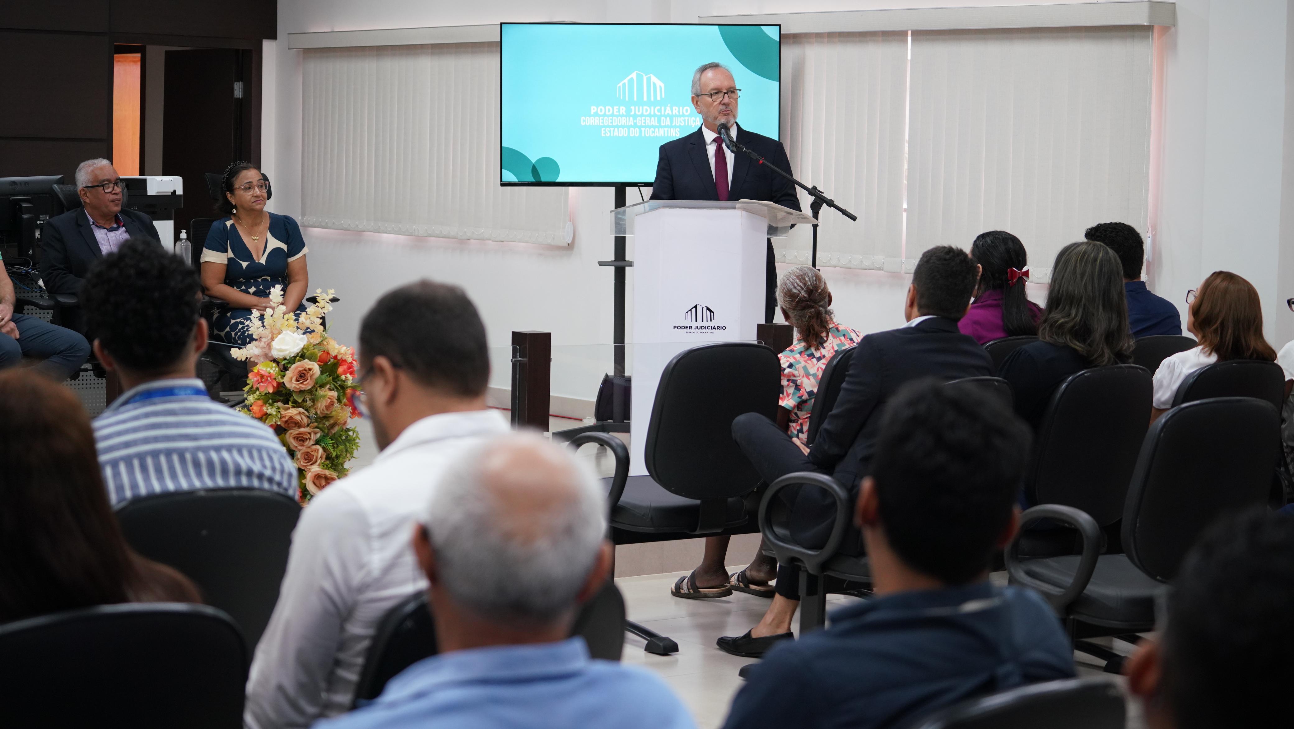 Homem branco de terno escuro fala em púlpito para servidores(as) em auditório de paredes brancas e persiana bege. Ao fundo, um telão exibe imagem com fundo azul claro