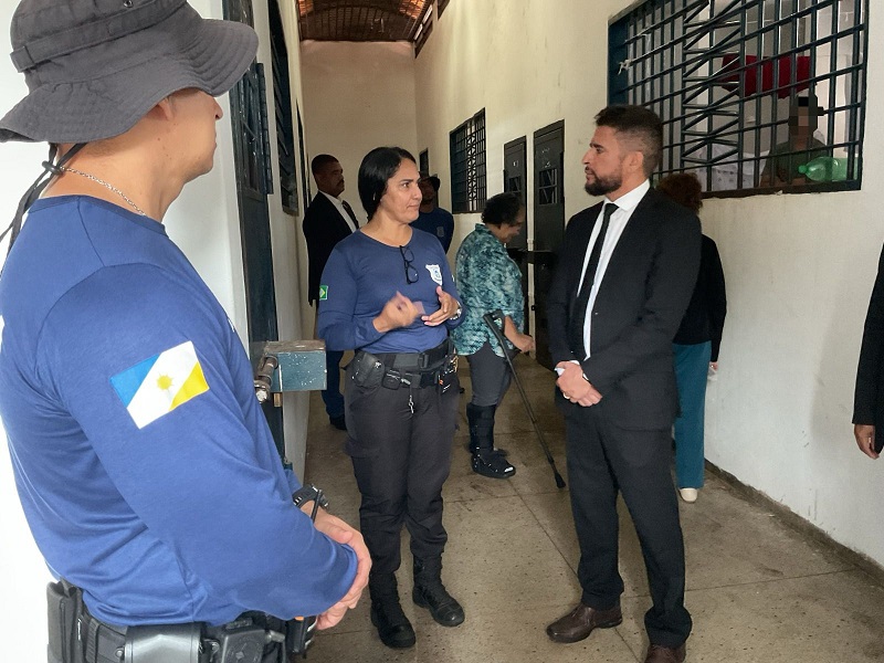 Homem de camisa azul e chapéu cinza observa mulher de camisa azul e calça escura, de cabelos presos, conversando com homem de terno escuro e camisa branca em ala de unidade de atendimento de adolescente em conflito com a lei