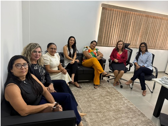 Foto aberta com sete mulheres sentadas em uma sala, sendo uma delas a juíza Cirlene, que está de blusa e blazer vermelhos e saia preta