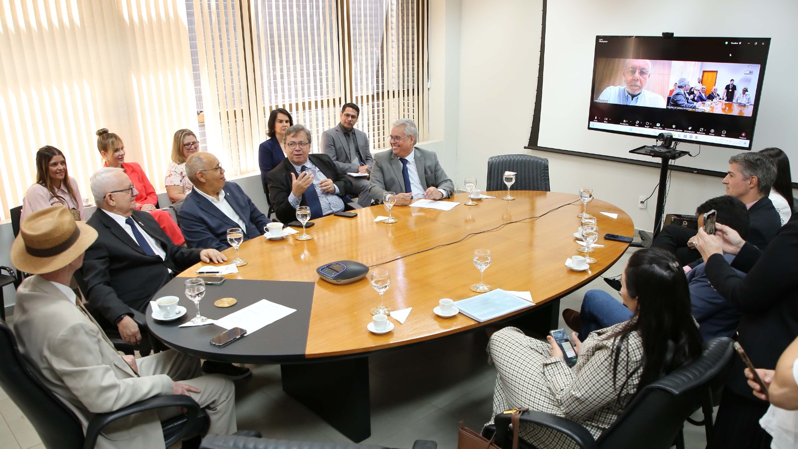 Na imagem aparecem 14 pessoas reunidas em torno de uma grande mesa oval de madeira em uma sala de reunião. Um dos participantes, um homem de terno escuro e camisa azul, gesticulam enquanto fala, e os demais escutam atentamente, alguns sorrindo. No fundo da sala, há uma tela de projeção mostrando uma videoconferência, onde um homem de óculos está aparecendo.