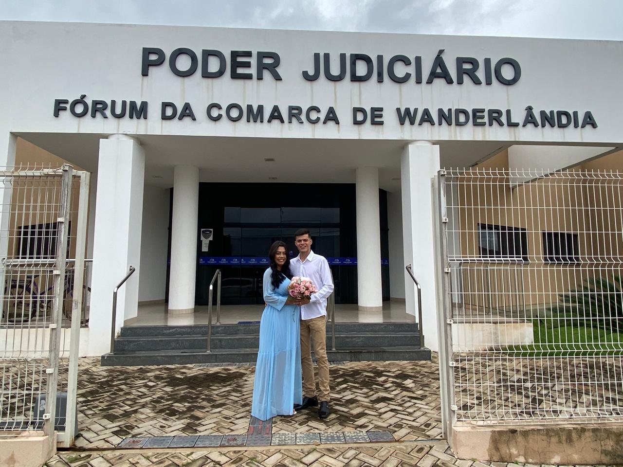Na imagem aparece um casal abraçado em frente ao fórum da comarca de Wanderlândia. O mulher está com um vestido longo na cor azul e o homem com calça marrom e camisa branca. Eles seguram um buquê de flores rosas. 