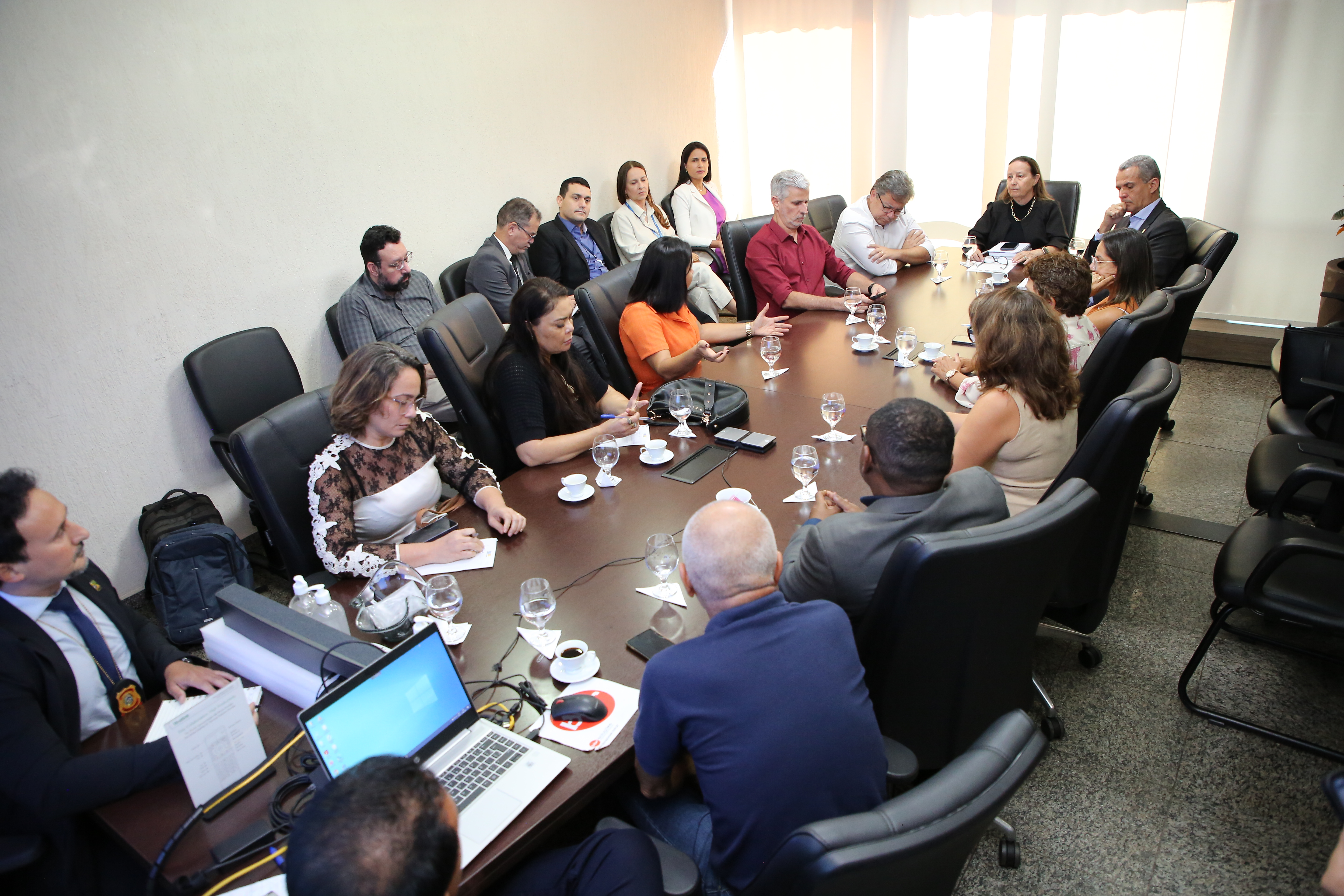 Fotografia aberta de uma reunião de grupo em torno de uma mesa grande, com várias pessoas sentadas em cadeiras, em uma sala.