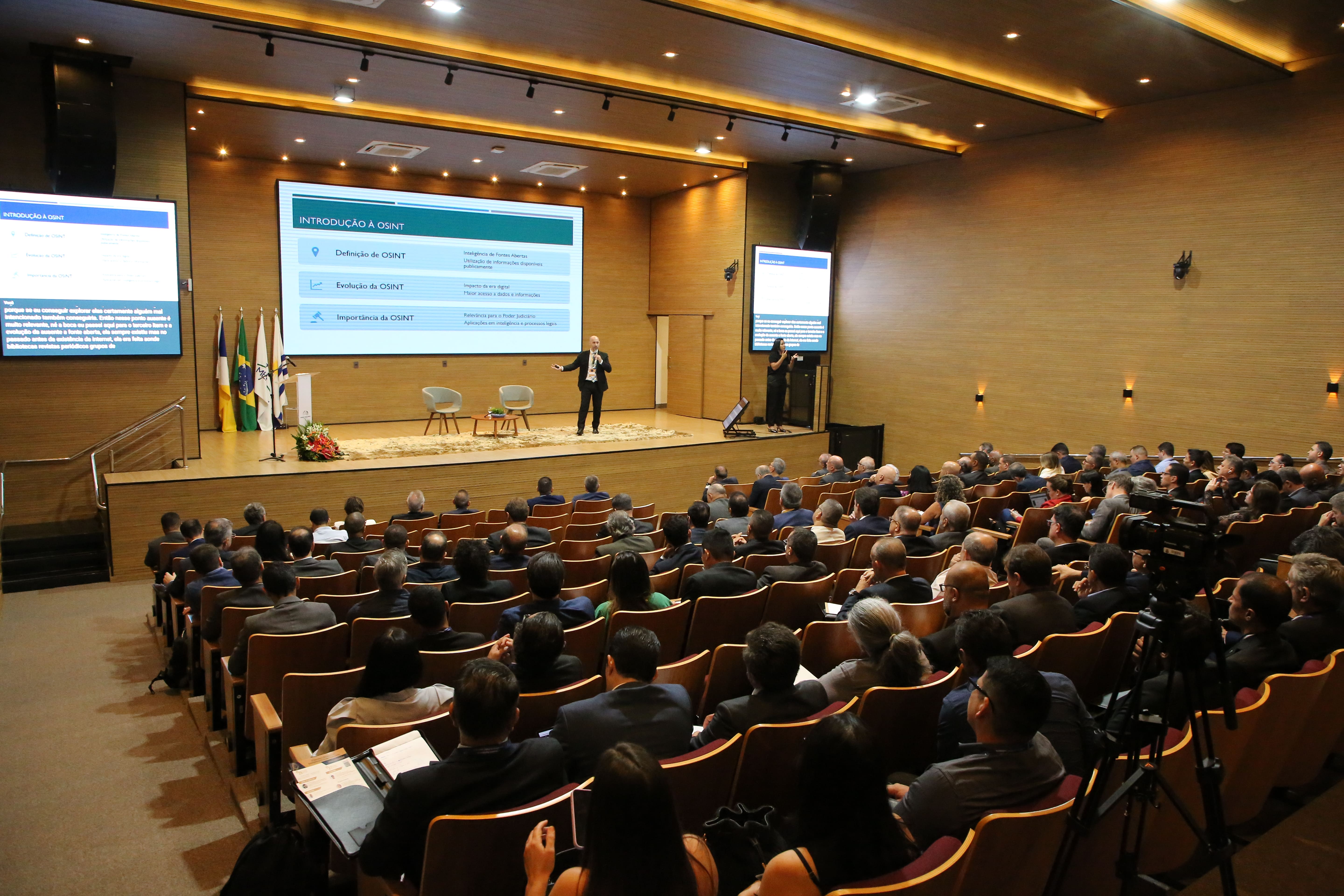 Imagem mostra um amplo auditório com muitos participantes assistindo a uma palestra. Há várias telas grandes exibindo informações na área do palco. O espaço é bem iluminado e as pessoas estão em postura atenta, demonstrando interesse pela palestra.