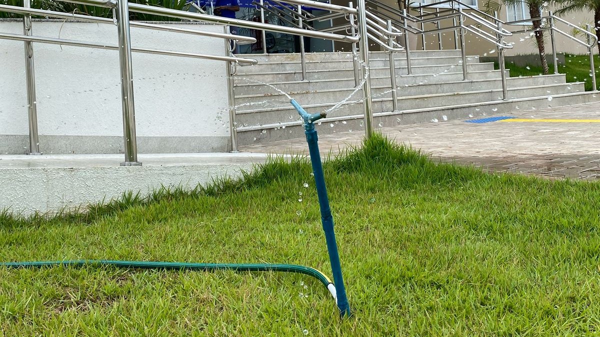 Foto de uma mangueira irrigando um jardim com uma grama bem verde; ao fundo, a escadaria e rampas de acesso ao prédio do Fórum