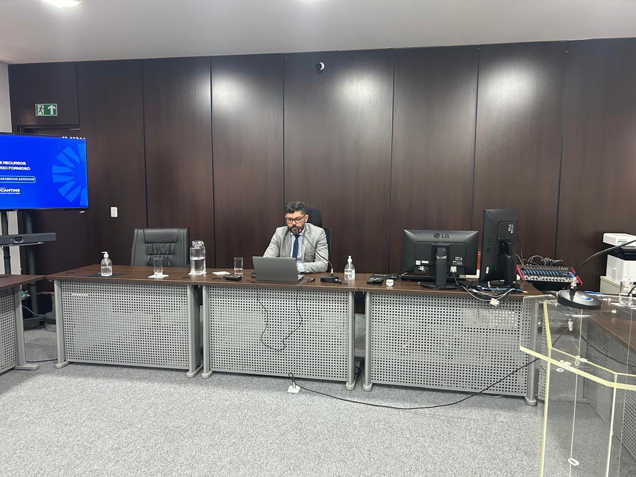 A fotografia mostra o juiz Wellington Magalhães sentado em frente a um computador na sala de audiência com uma mesa e monitores.