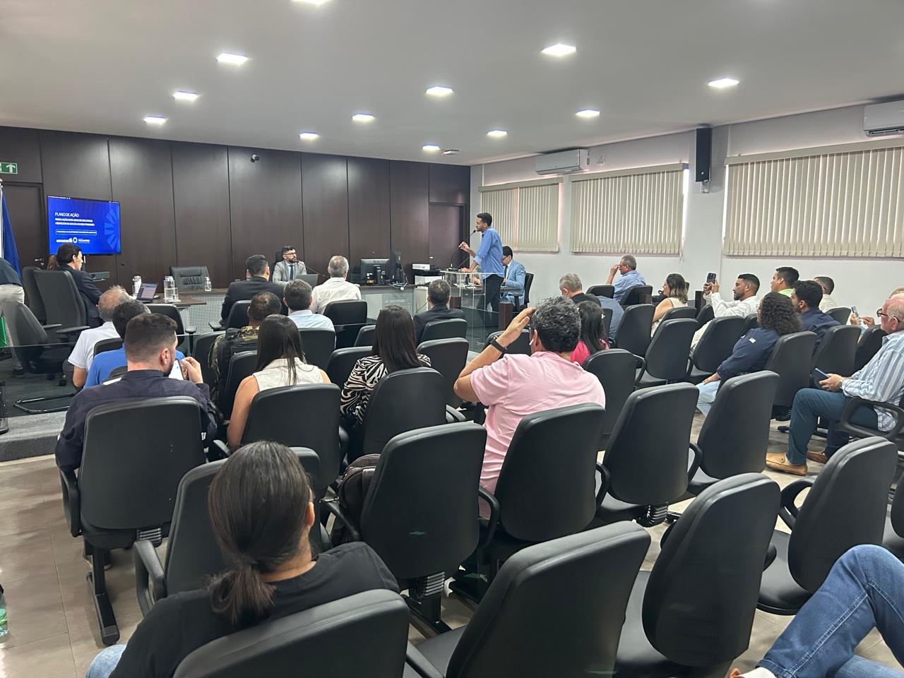 A fotografia mostra uma grande sala de reuniões com várias pessoas sentadas em fileiras de cadeiras pretas. Há uma plataforma elevada com algumas pessoas em pé ou sentadas.