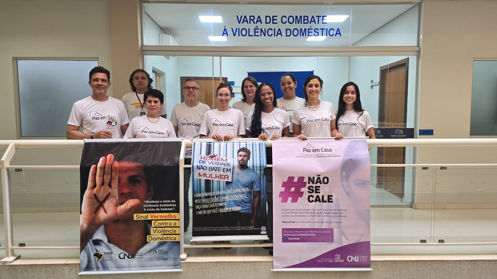 A fotografia mostra um grupo de pessoas com camisetas brancas da Semana pela Paz em Casa, com cartazes em apoio à campanha contra a violência doméstica. As pessoas estão posando em frente à sala da Vara de Combate à Violência Doméstica.