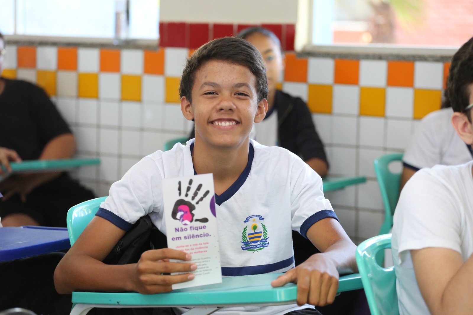A foto mostra um jovem sorridente, em uma sala de aula, segura um panfleto sobre não ficar em silêncio.