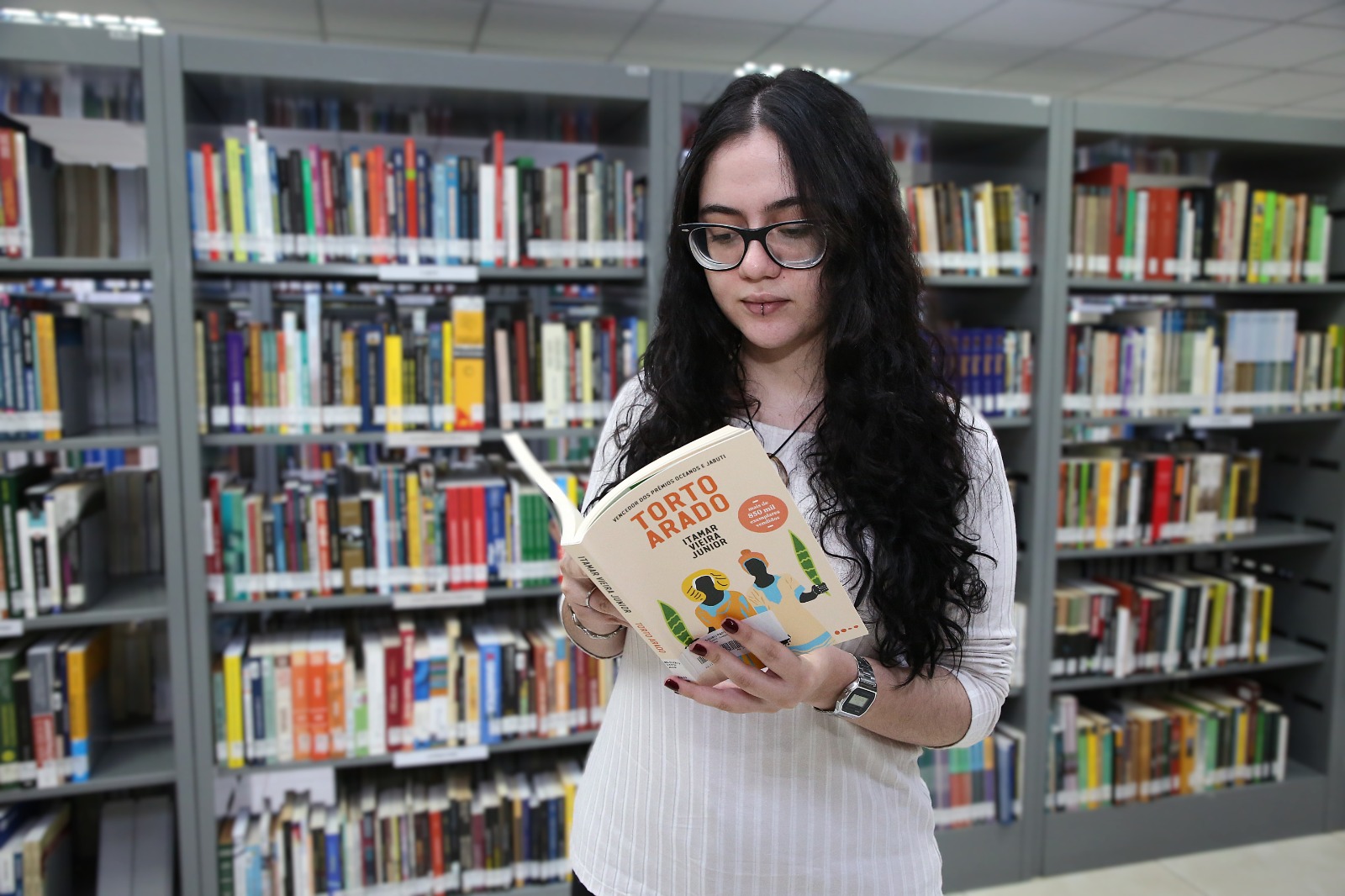 A imagem mostra uma jovem lendo um livro, em foco principal, e ao fundo tem várias  prateleiras com muitos livros de diferentes cores e formatos, criando uma atmosfera de estudo e aprendizado.