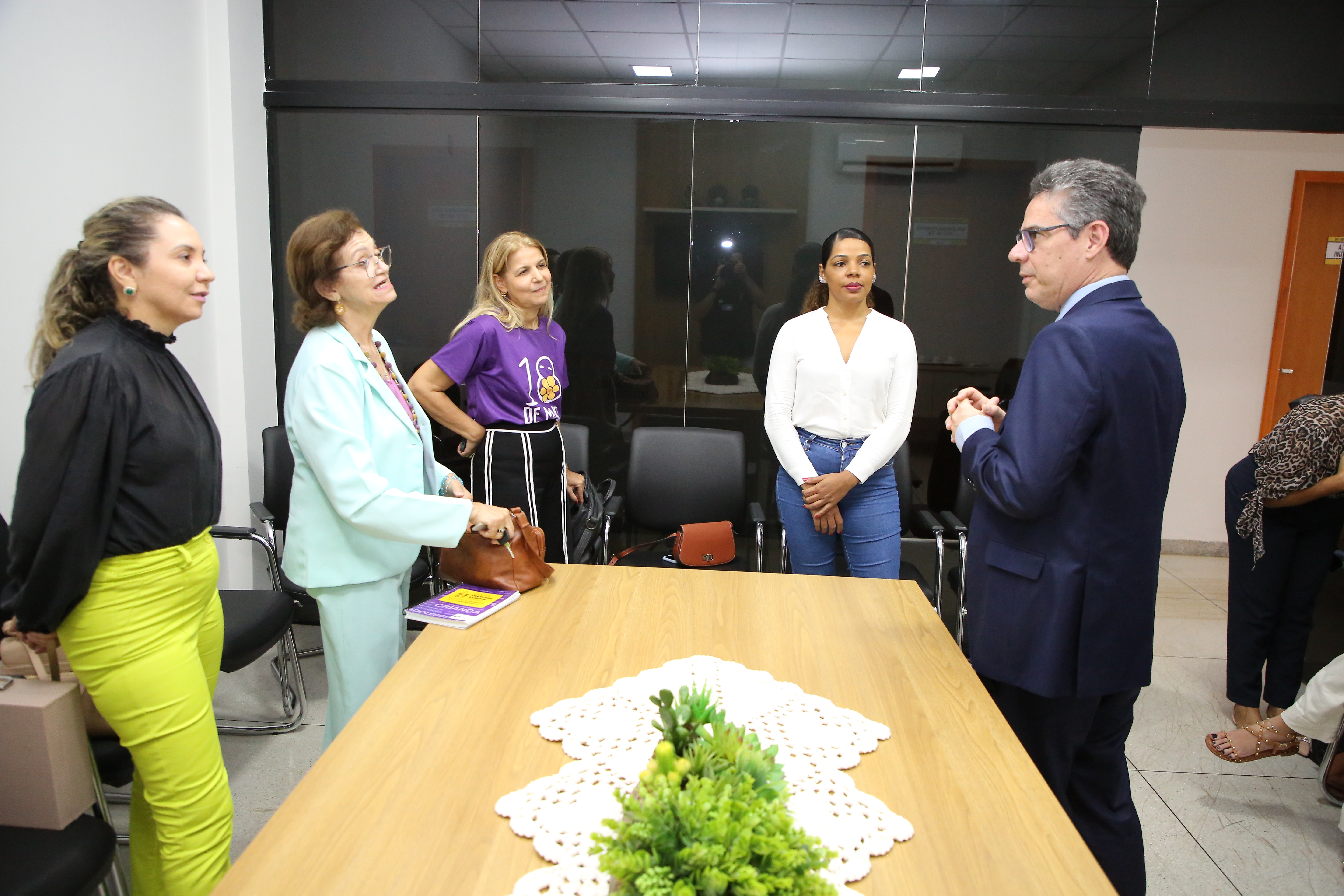Na imagem aparecem cinco pessoas, 4 mulheres e 1 homem, em volta de uma mesa de madeira, em uma sala de reunião. Elas estão conversando sobre a lei da escuta protegida.