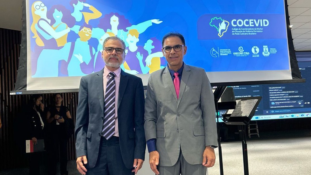 Dois homens de óculos de grau e terno posam em frente a um telão que projeta uma imagem em tons azuis sobre evento voltado ao combate à violência doméstica