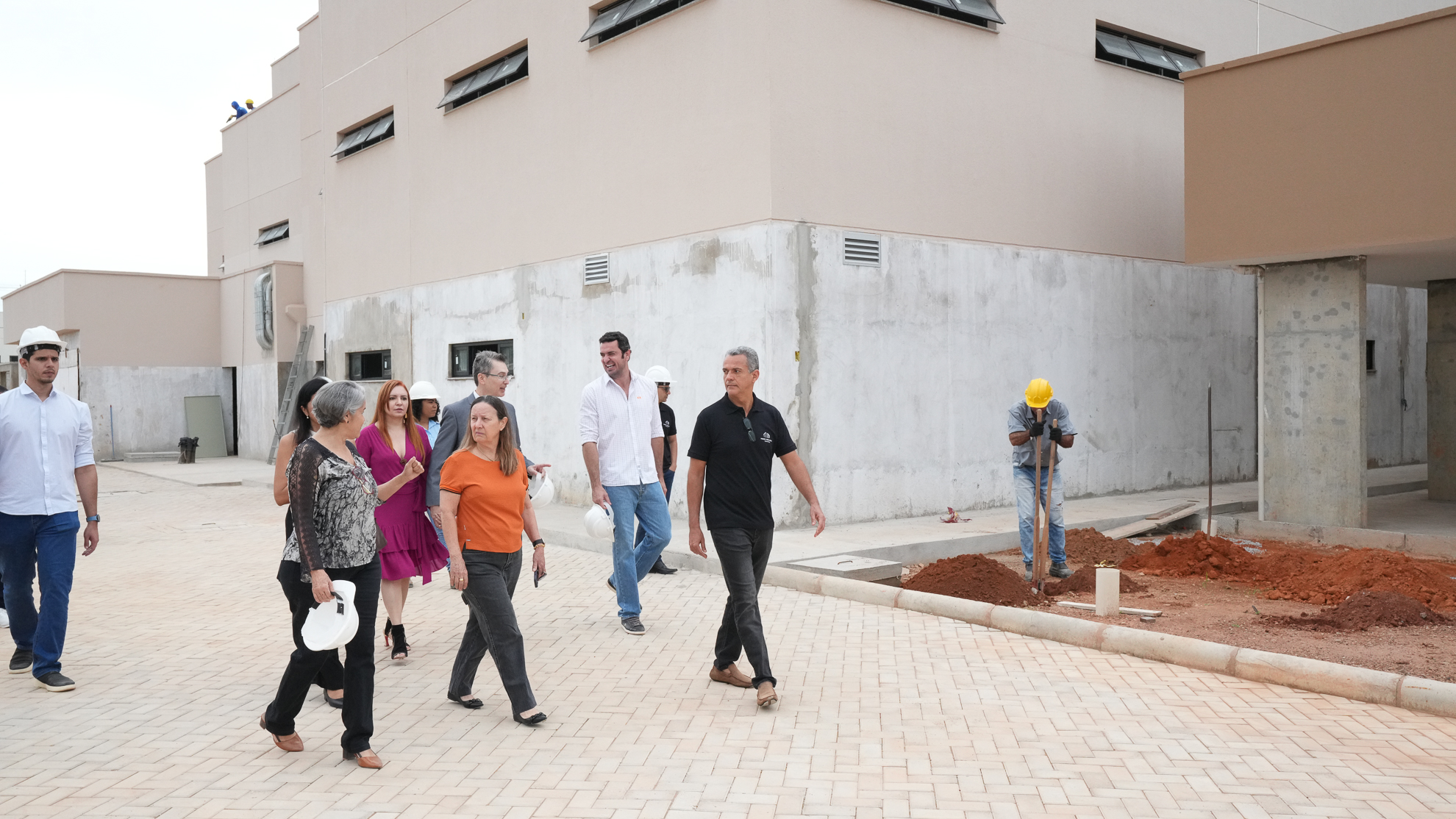 A imagem mostra um grupo de pessoas caminhando em um local de construção. As pessoas estão distribuídas ao longo de um calçamento. Em um canto da imagem tem um trabalhador que está cavando um buraco.