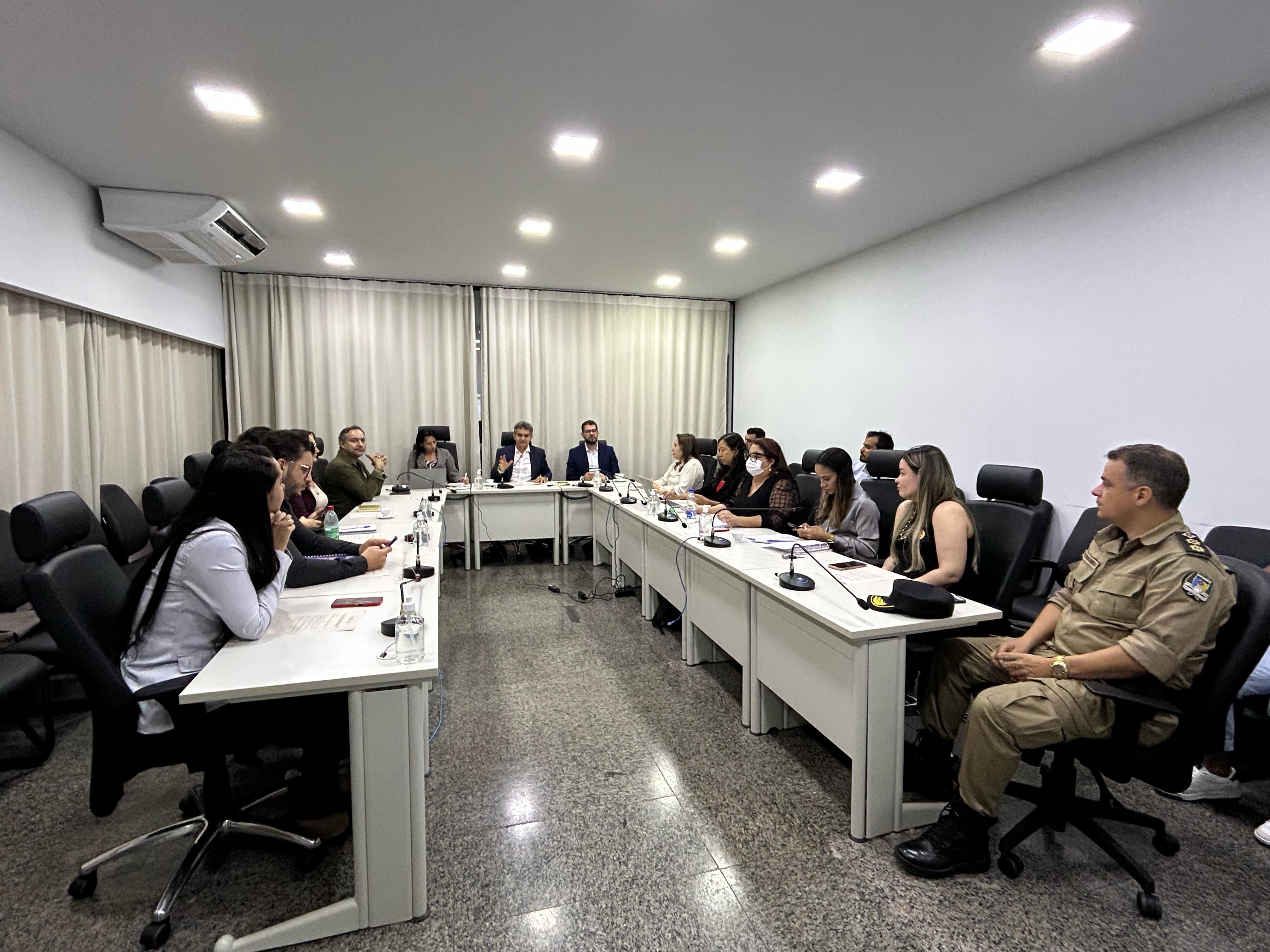 Homens e mulheres sentados em mesa branca em formato U, em sala de paredes brancas e persiana bege