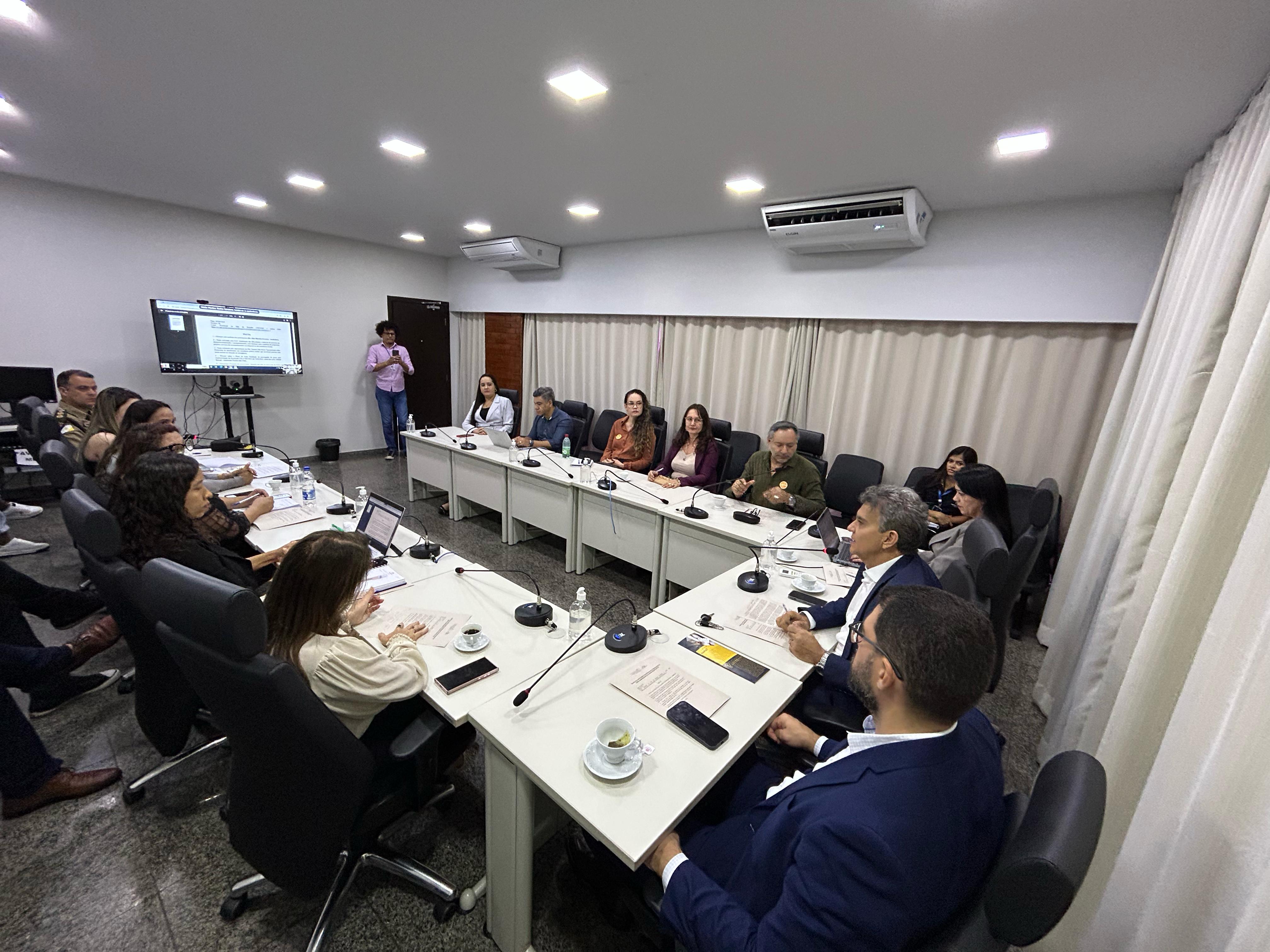 Homens e mulheres sentados em mesa branca em formato U, em sala de paredes brancas e persiana bege