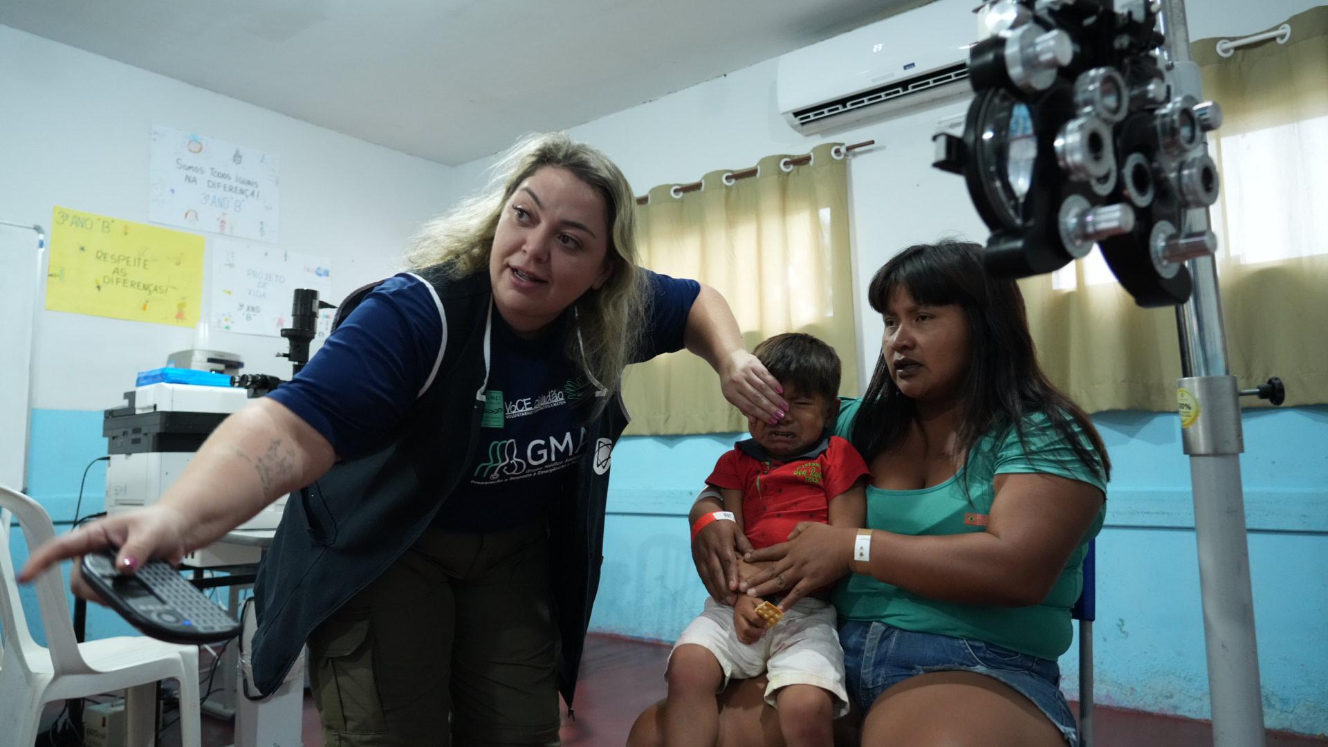 Mulher indígena sentada com filho bebê no colo durante consulta oftalmológica com mulher branca, loura que tapa uma das vistas do bebê com uma mão e aponta para a frente com a outra 