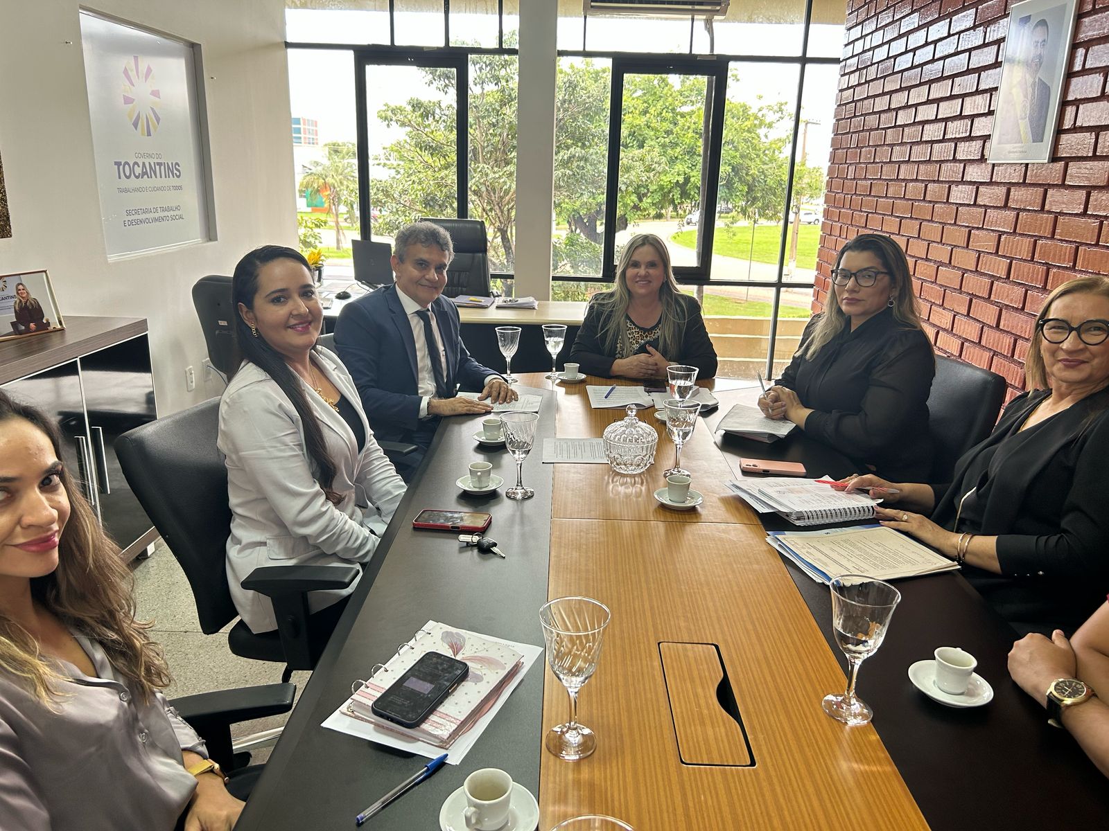 A imagem mostra um grupo de pessoas em uma sala de reunião, sentadas em torno de uma grande mesa retangular. 