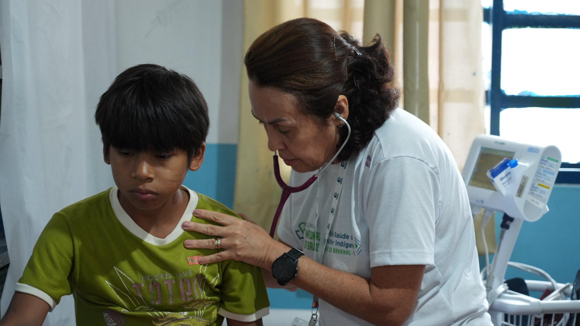 Foto de mulher de cabelos escuros na altura dos ombros, profissional de saúde de camisa branca, usando estetoscópio para ascultar pulmão de menino indígena que está sentado usando camiseta verde. Eles estão numa sala de parede branca com azul e cortina bege