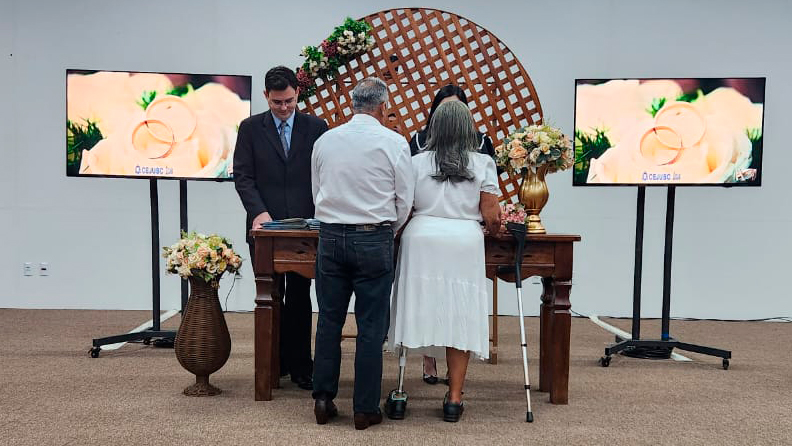 Imagem de casal de costas durante celebração de casamento civil. Tem um juiz uma juíza segurando documentos em cima de uma mesa de madeira. Ao fundo, um adorno de madeira de treliça e aos lados, telas projetam flores brancas